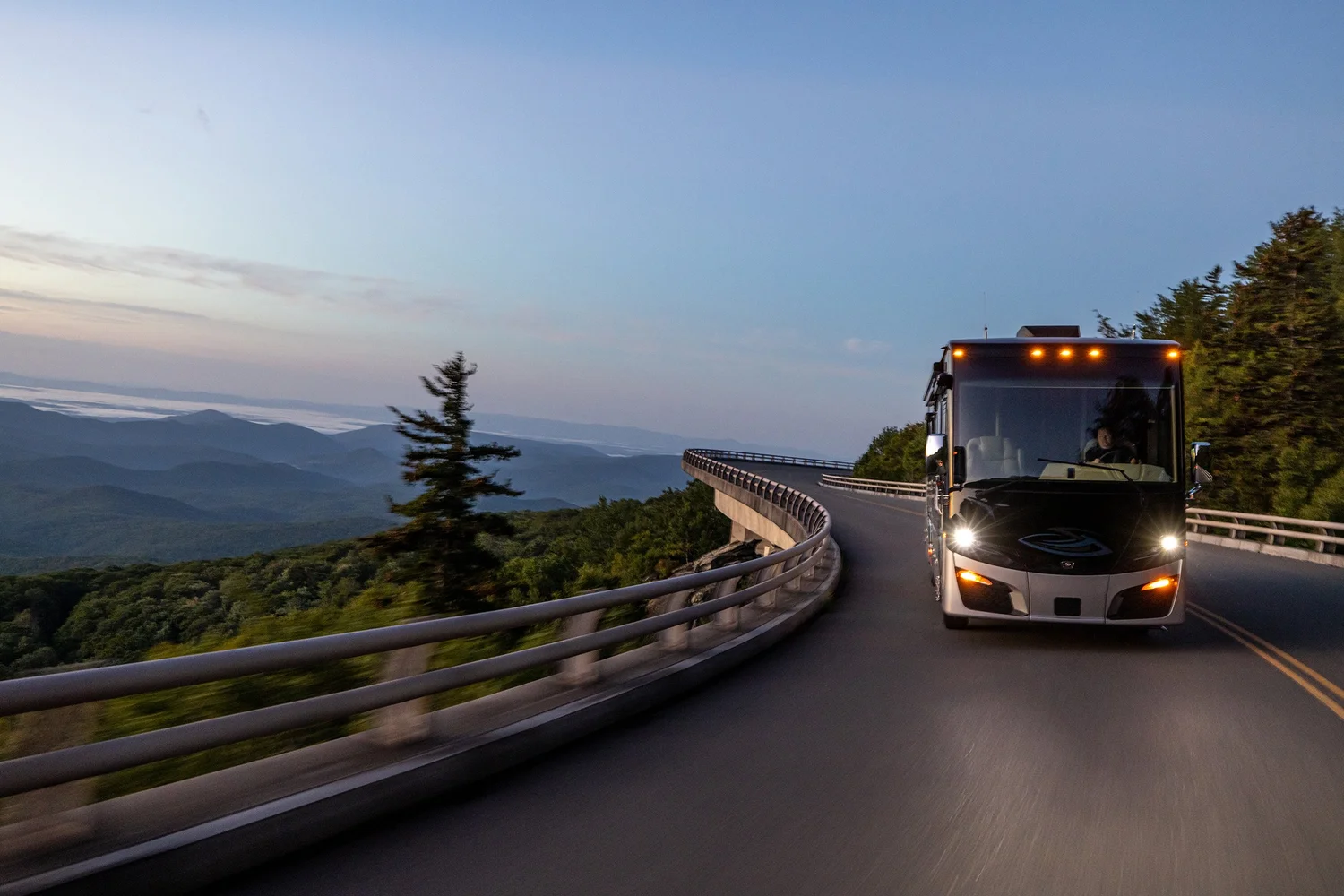 A modern bus traveling on a winding mountain road at dusk, with a scenic view of hills and trees in the background.