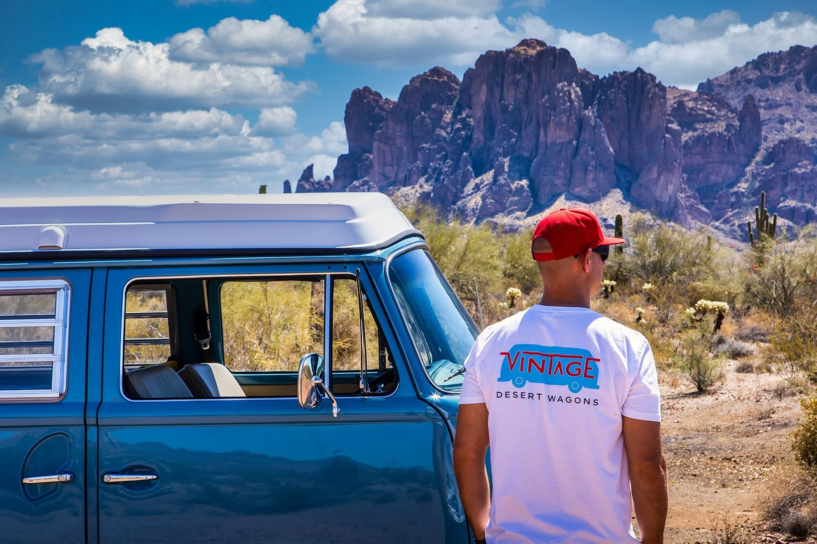 A man wearing a white T-shirt with the words ‘VINTAGE DESERT WAGONS’ and a red cap stands near a vintage blue camper van in a desert landscape with cacti and rocky mountains in the background.
