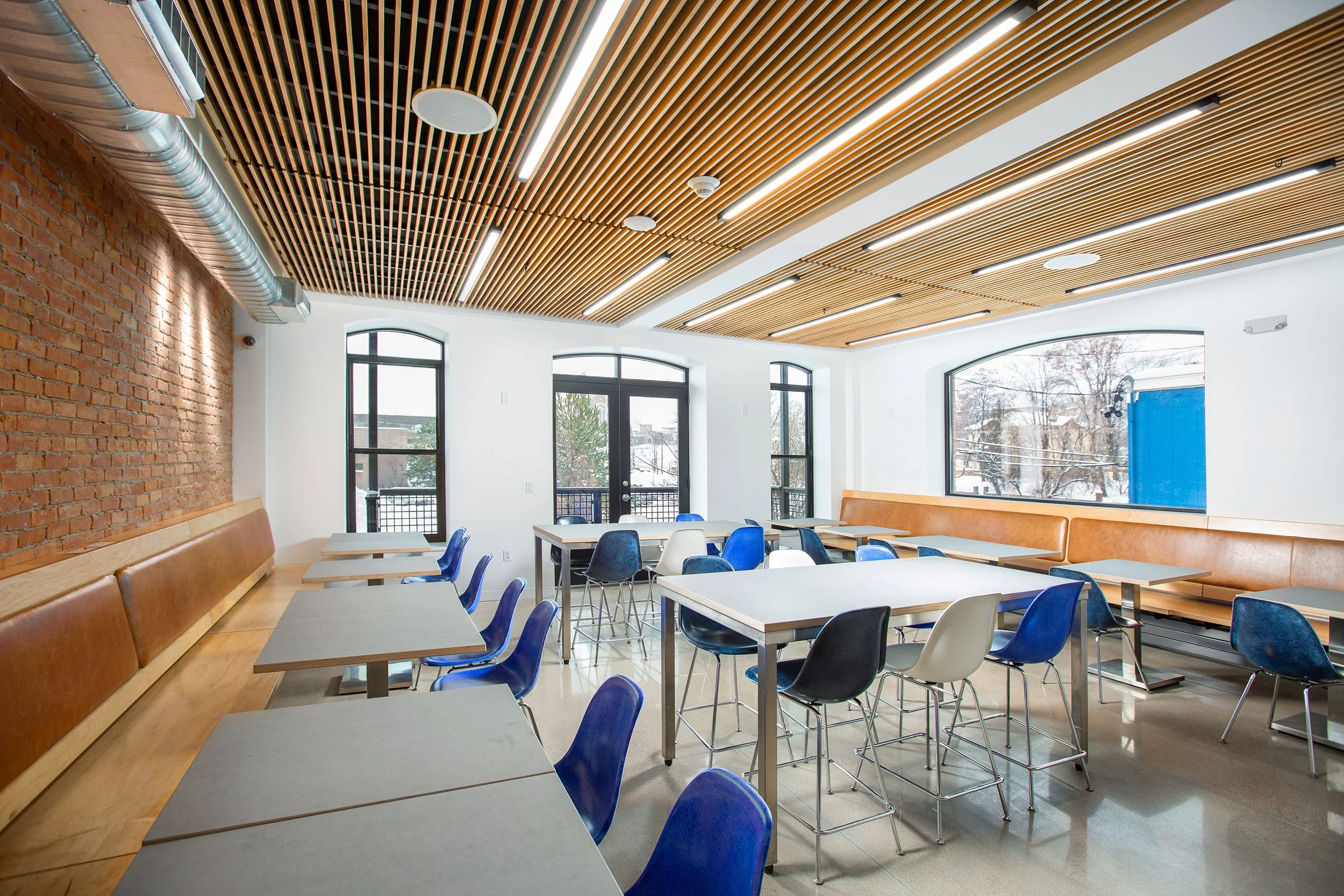Modern cafeteria with exposed brick wall, wooden slat ceiling, large windows, and a mix of blue, white, and black chairs around light-colored tables.