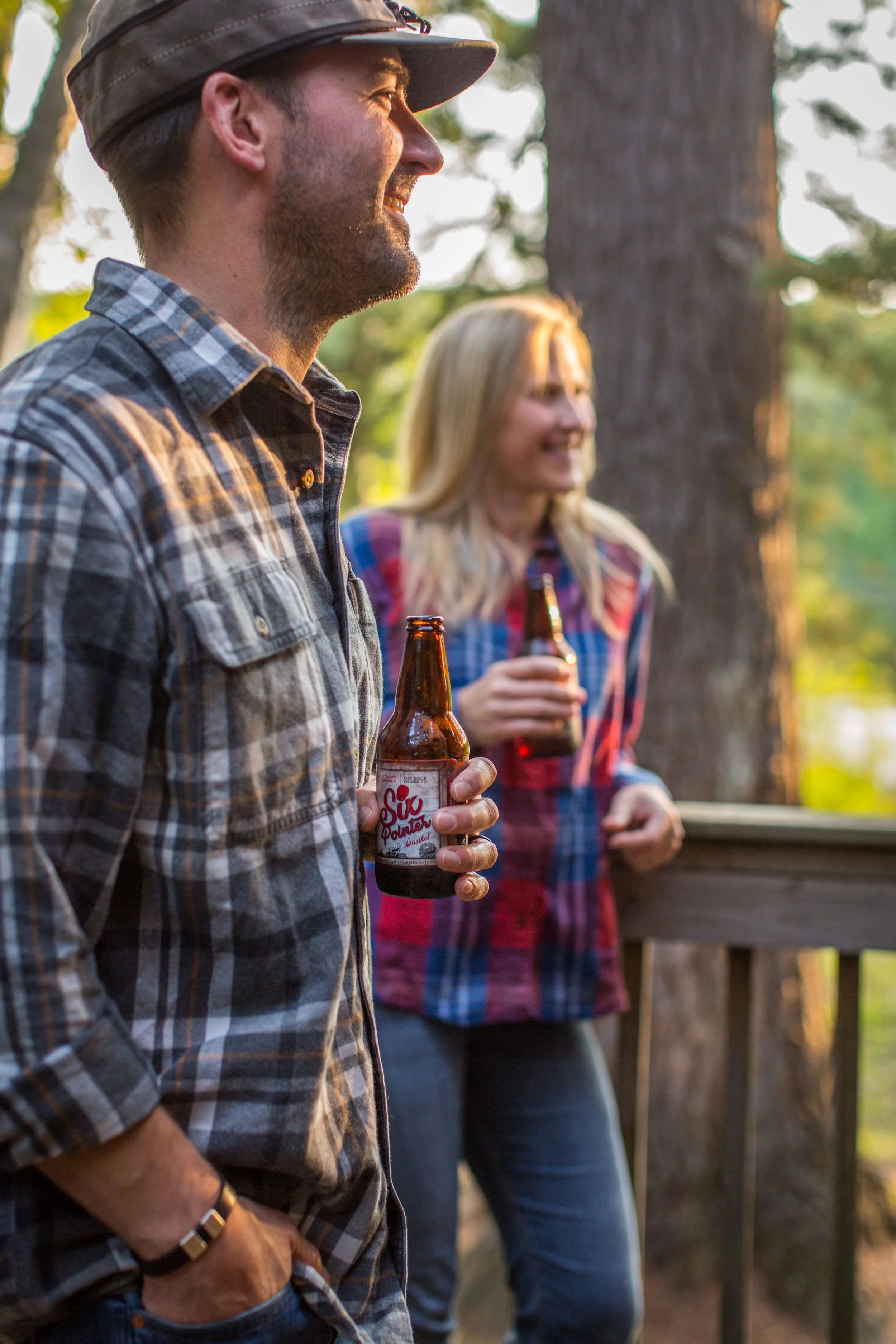 A man and a woman are outdoors in a wooded area, holding bottles of beer and smiling, enjoying a casual social gathering.