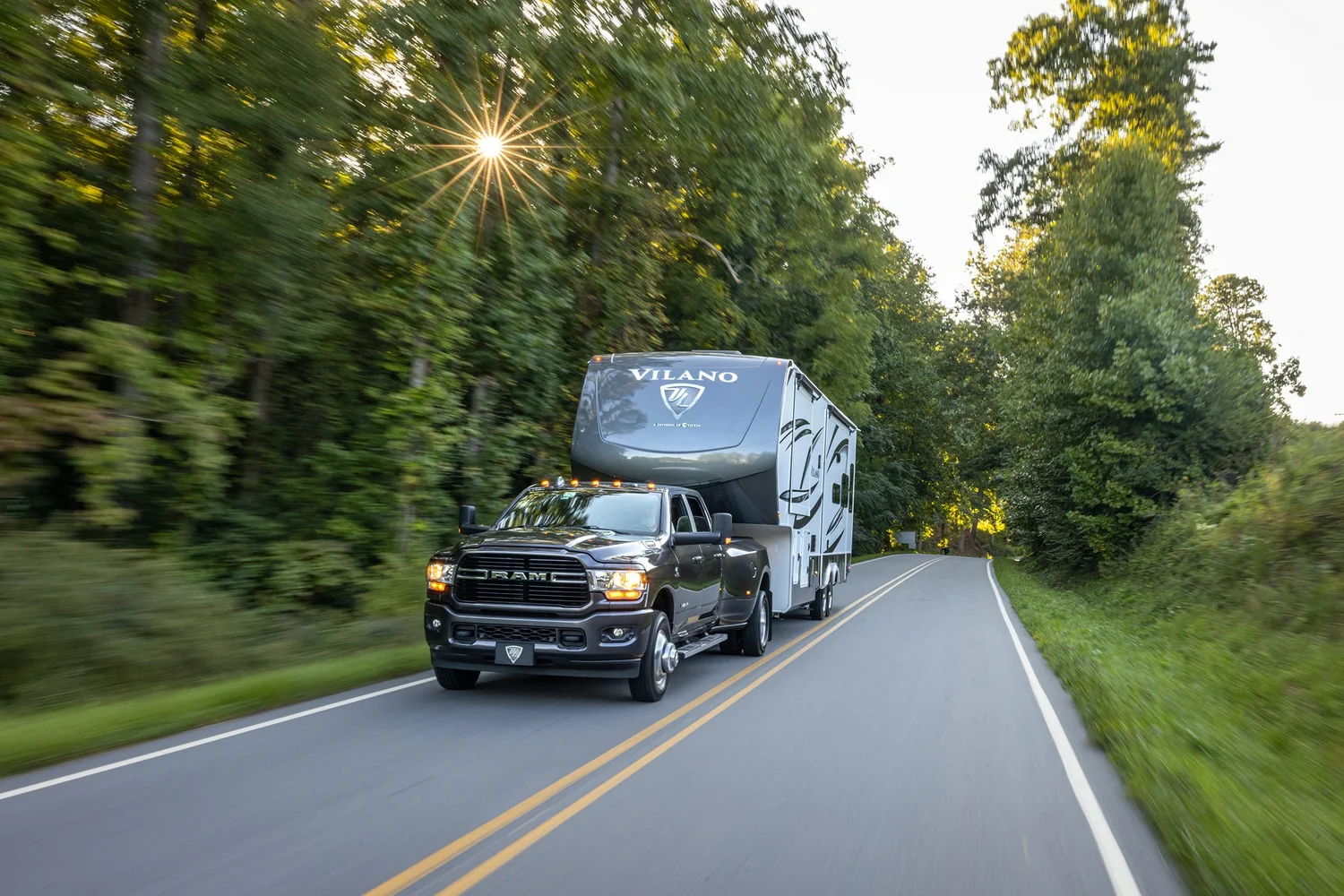 A black RAM pickup truck towing a Vilano travel trailer on a winding road surrounded by green trees, with the sun shining through the foliage.