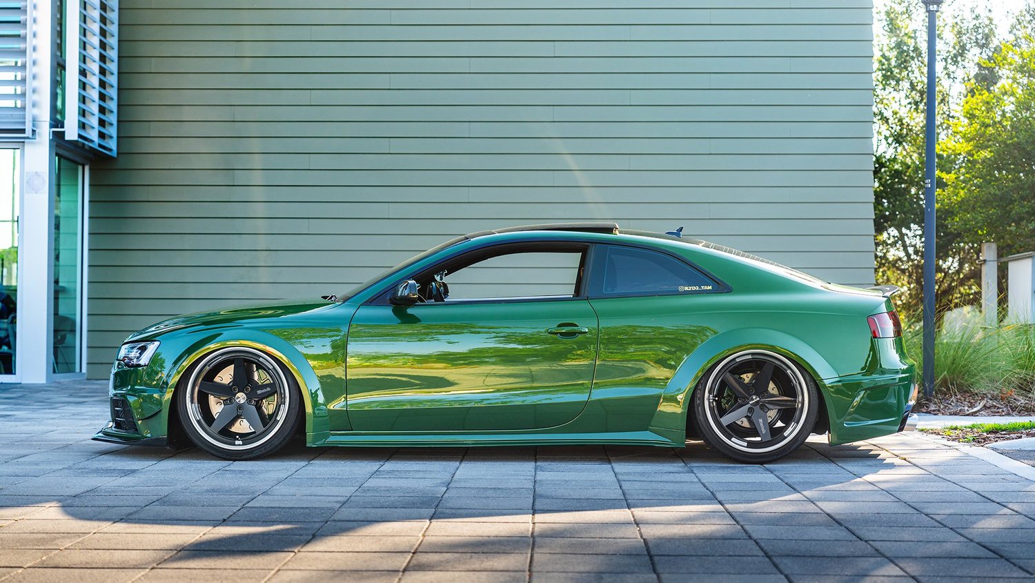 Green modified sports car parked on paved ground next to a building with gray siding.