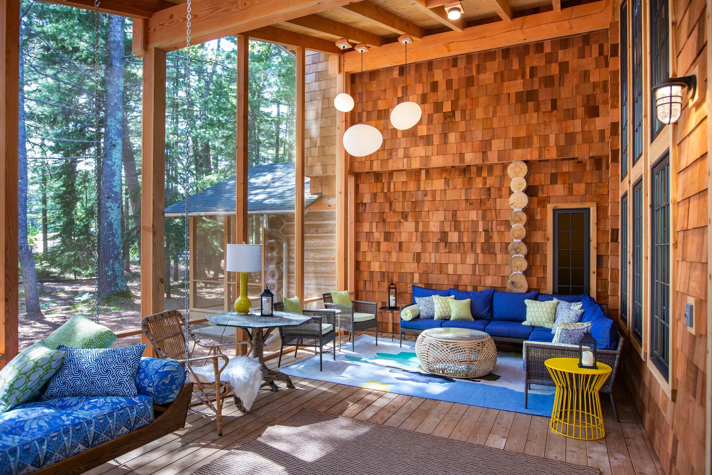 Sunroom with wood-paneled walls, large windows, outdoor forest view, hanging white globe lights, blue sectional sofa with green and blue pillows, wicker chairs, colorful area rug, and various decorative items.