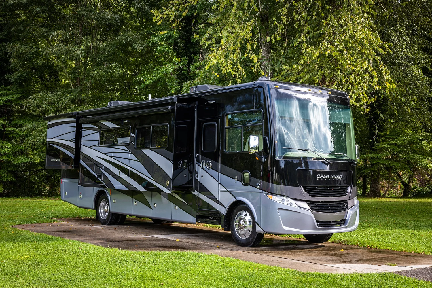 Black and gray recreational vehicle (RV) parked on a concrete pad in a grassy area surrounded by trees.