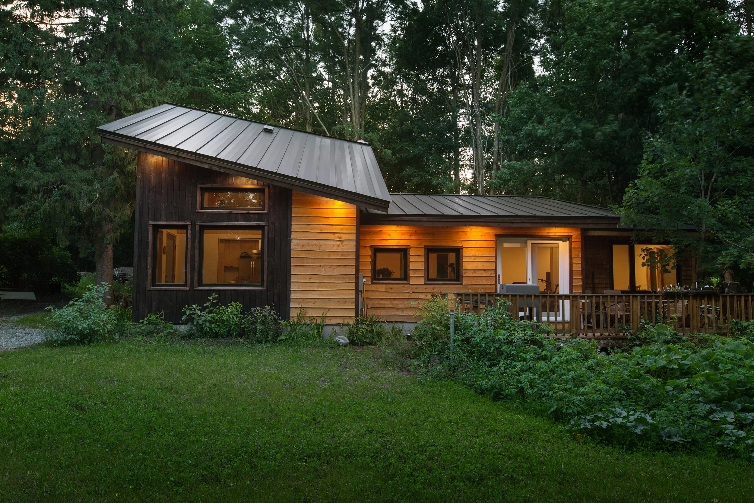 A modern house with a metal roof and wooden siding, illuminated from inside, surrounded by green trees and a garden at dusk.