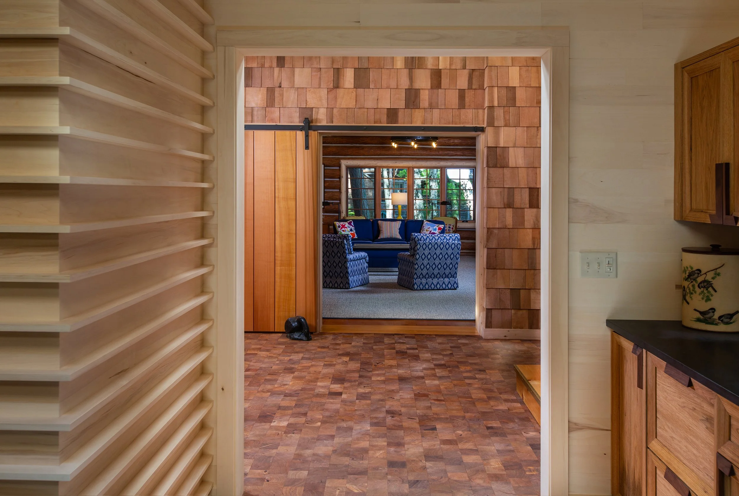 Interior view of a cozy living room with wooden walls, large windows, and a blue sofa with patterned pillows, seen through a doorway from a kitchen area with wooden cabinets and a countertop.