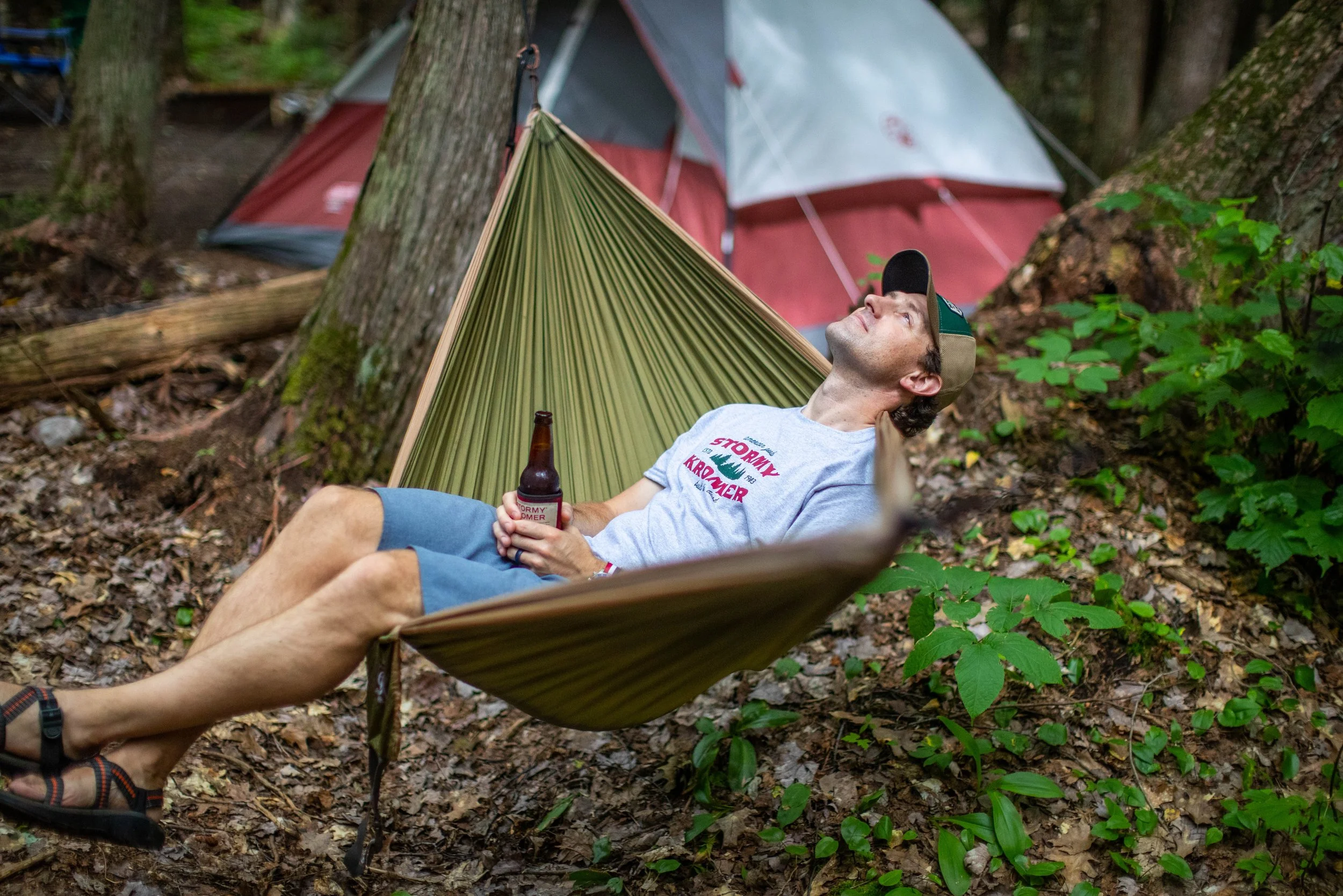A man relaxing in a hammock holding a beer, outdoors in a wooded area with camping tents in the background.