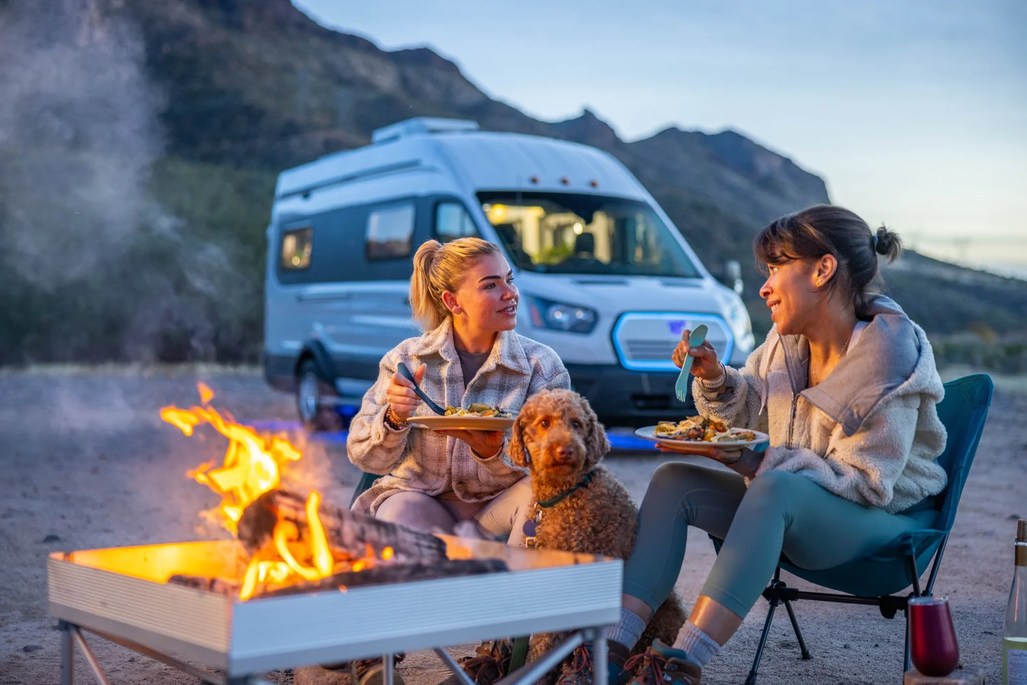 Two women sitting around a campfire enjoying dinner with a camper van in the background in a mountainous outdoor setting during dusk, with a dog sitting between them.
