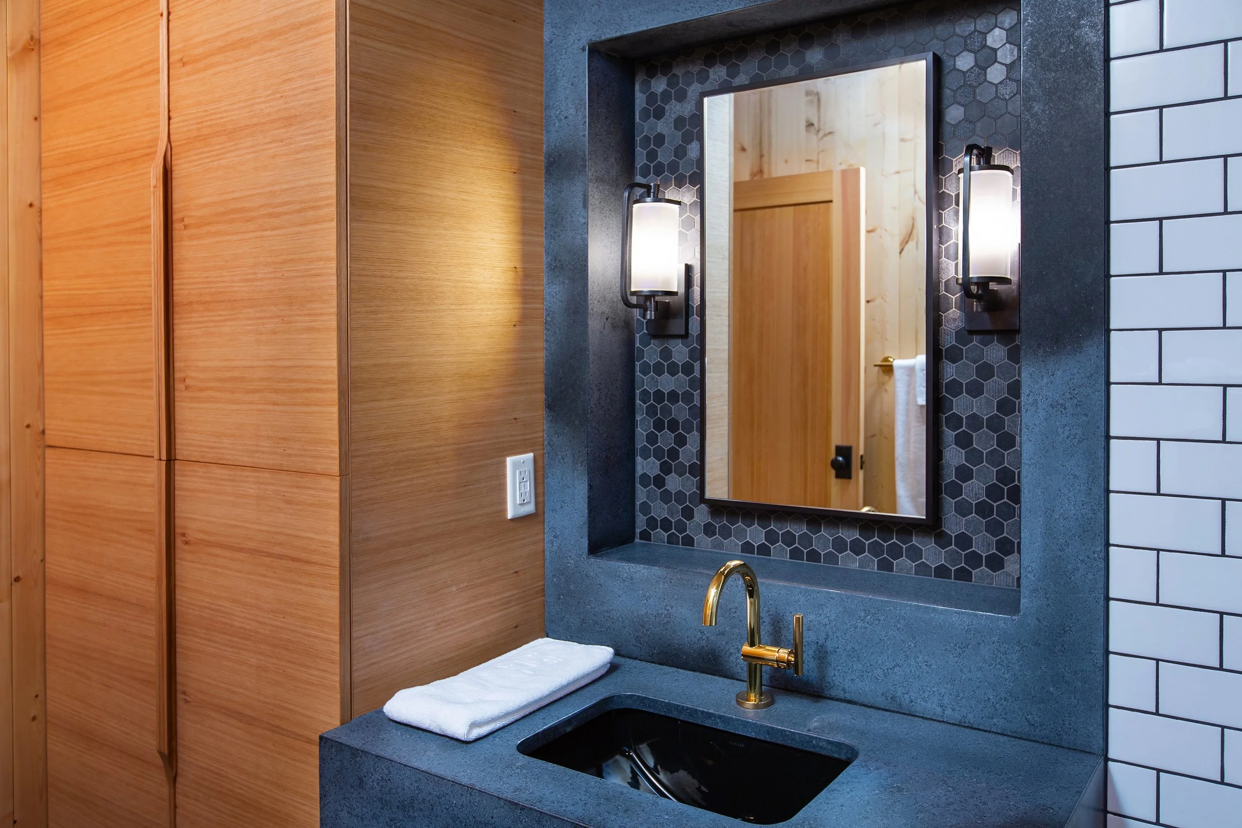 Bathroom with a wooden cabinet, a framed mirror, wall-mounted lights, a black sink, a gold faucet, and a folded towel on the countertop.