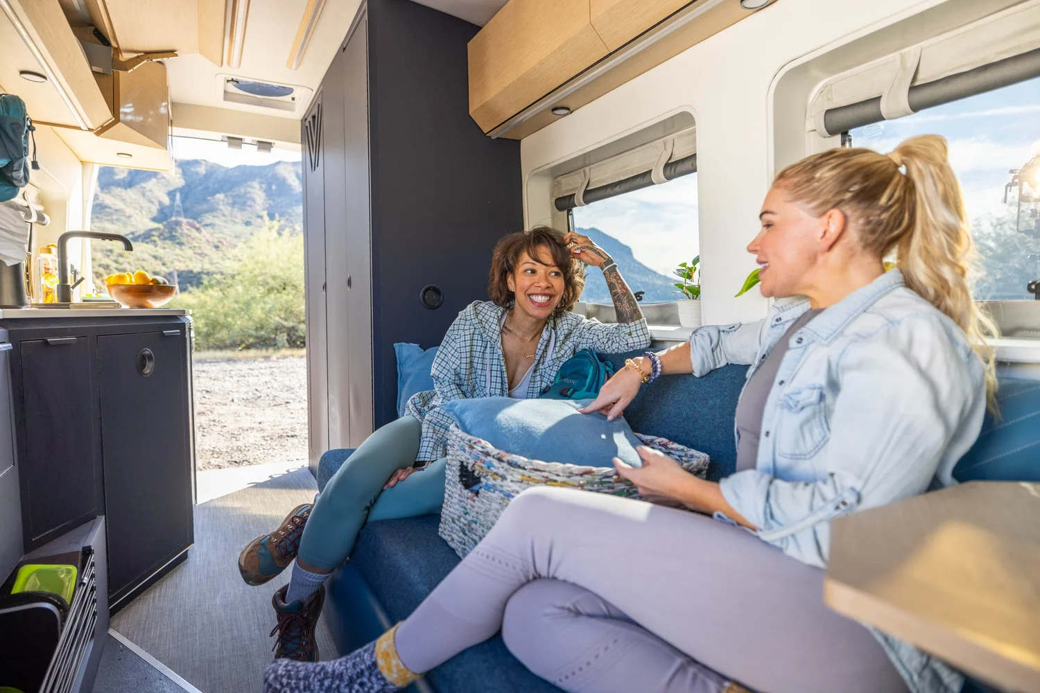 Two women sitting inside a camper van, talking and laughing with a scenic mountain view outside the window.