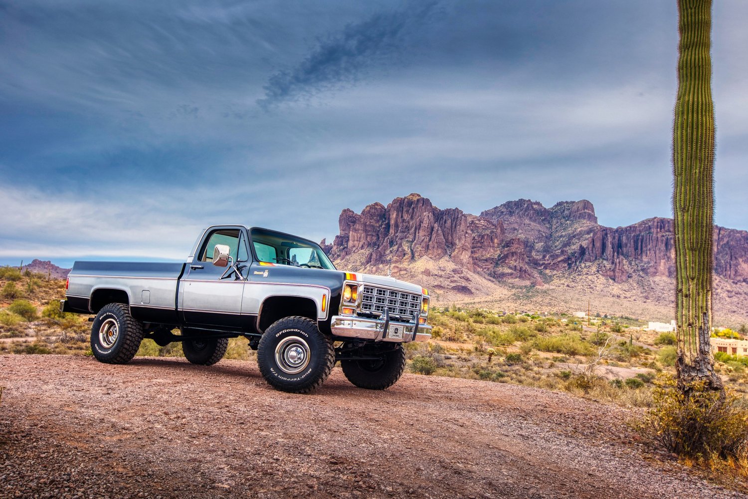 An off-road pickup truck parked on a dirt path in a desert landscape with mountain ranges in the background and a tall cactus on the right side.
