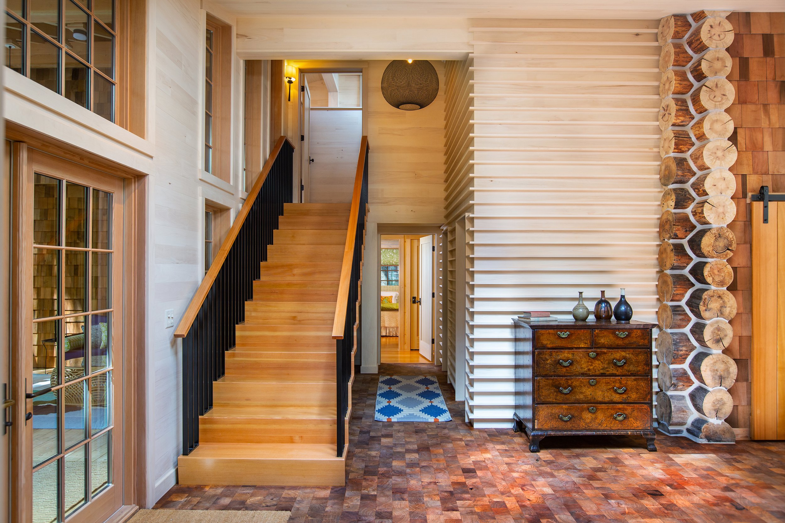 Interior of a modern house with wooden staircase, glass door, decorative wood logs, and a chest of drawers.