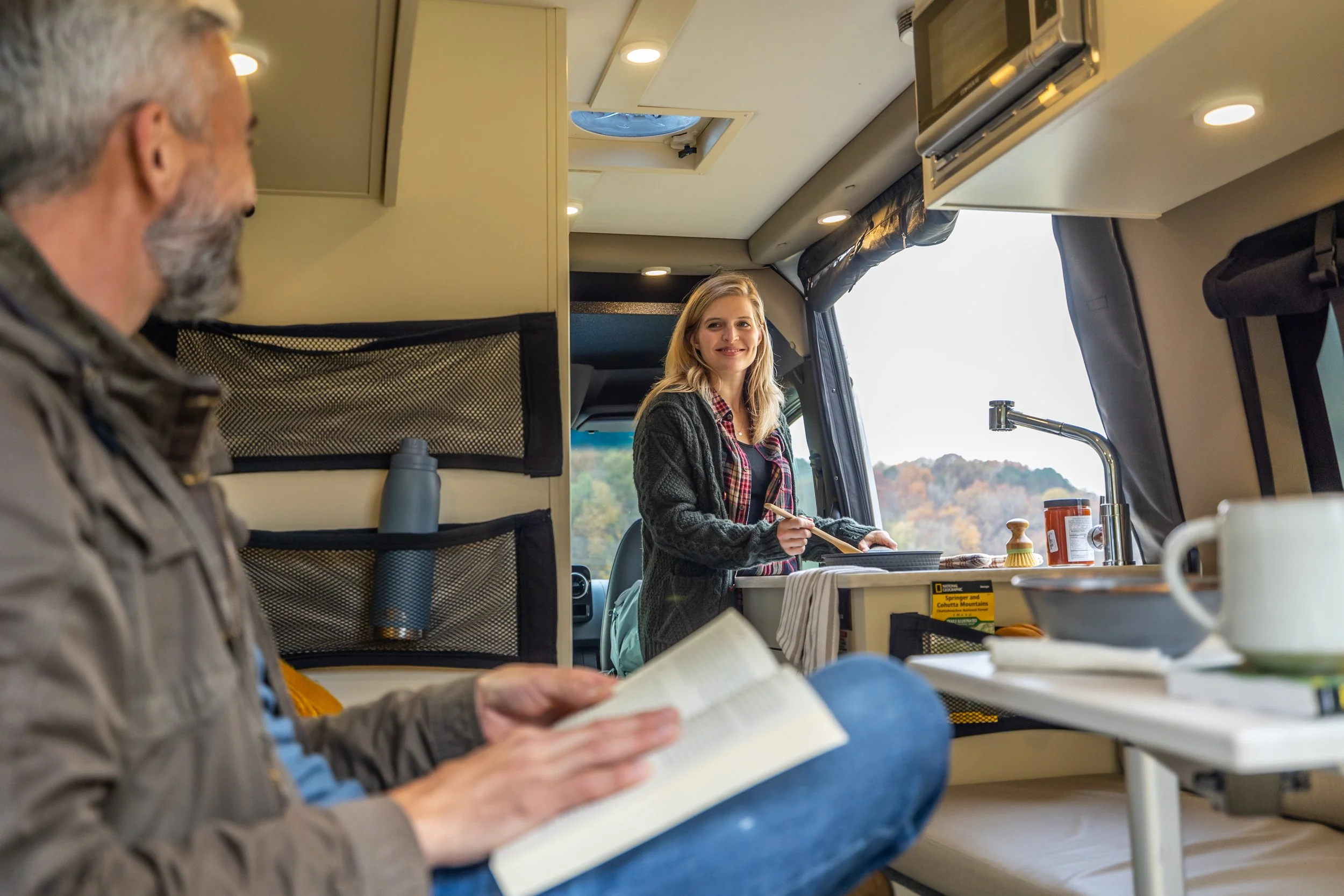A man reading a book inside a camper van with a woman cooking at the kitchen sink, with scenery of trees and hills outside the window.