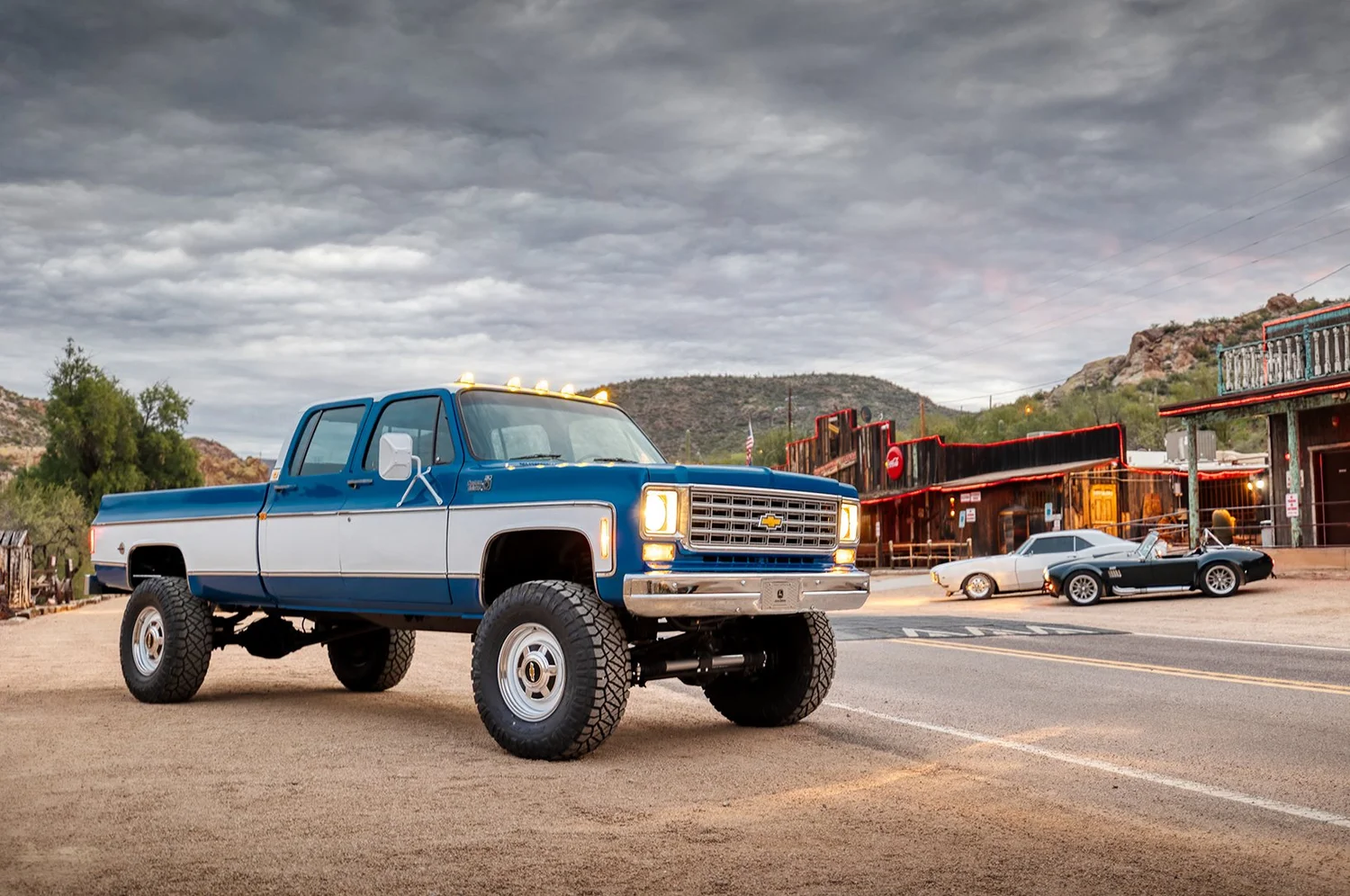 A blue and white vintage Chevrolet pickup truck parked on a dirt area in front of an old western style town with wooden buildings, and a couple of classic cars parked along the street under a cloudy sky.