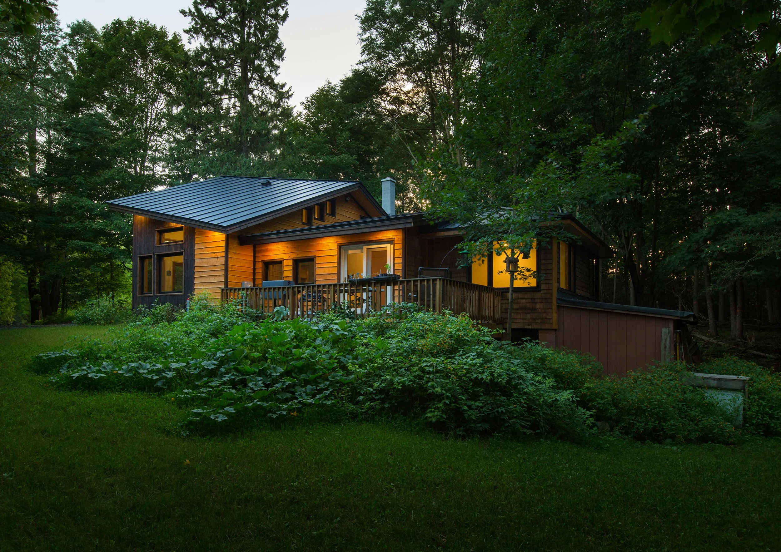 A cozy, wooden house with illuminated windows, surrounded by lush green trees and a garden at dusk.