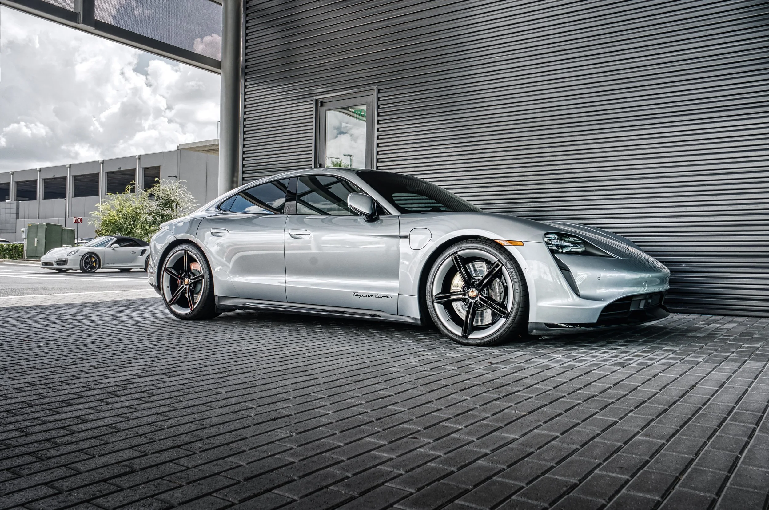A silver Porsche Taycan Turbo electric sports car parked outside a building with a black metal exterior, with a gray Porsche 911 Carrera in the background.