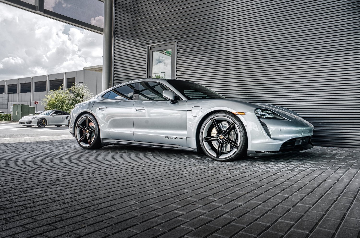 A silver Porsche Taycan Turbo electric sports car parked outside a building with a black metal exterior, with a gray Porsche 911 Carrera in the background.