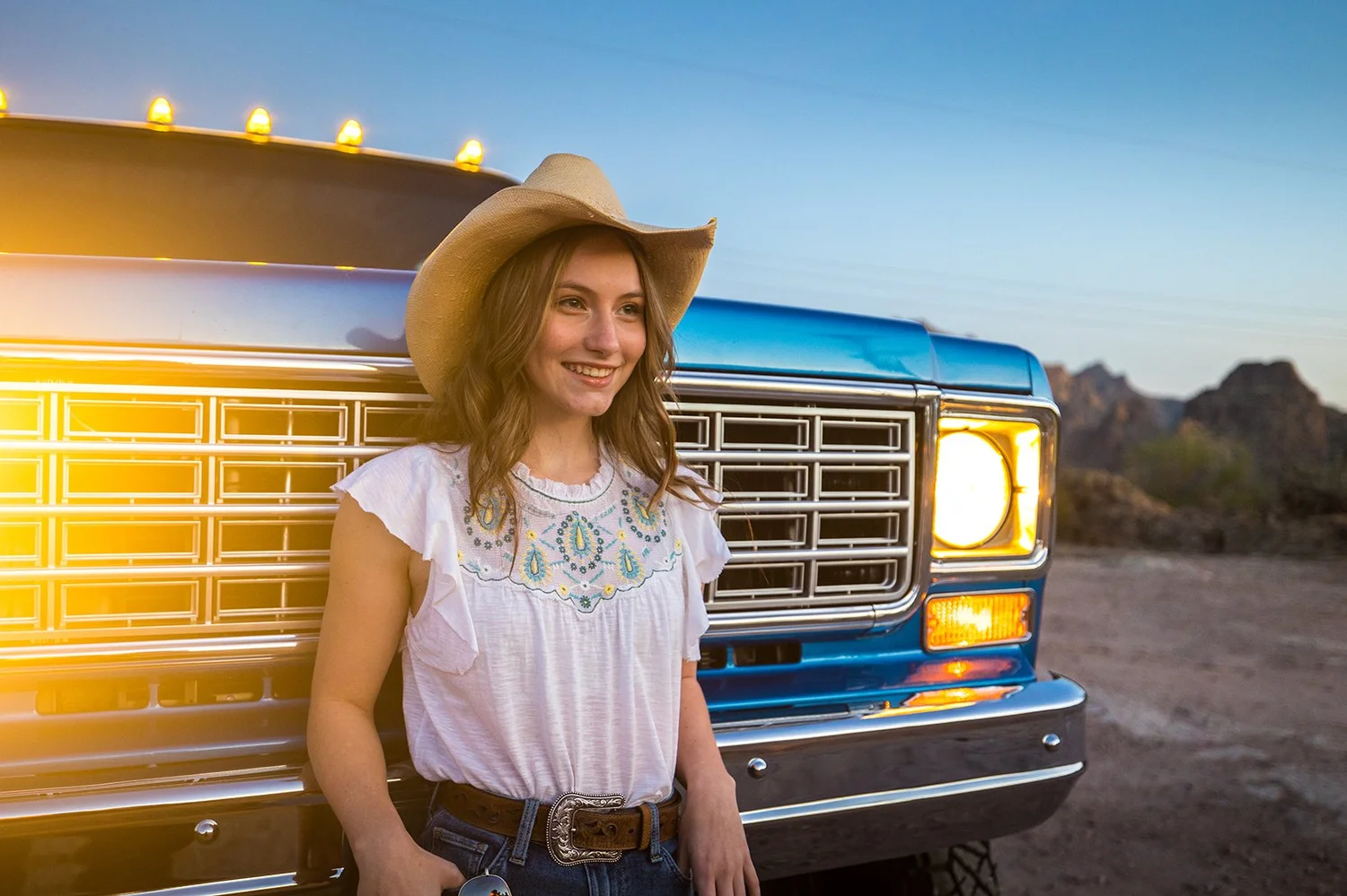 Young woman in a cowboy hat smiling, leaning against a vintage blue truck with mountains in the background during sunset.