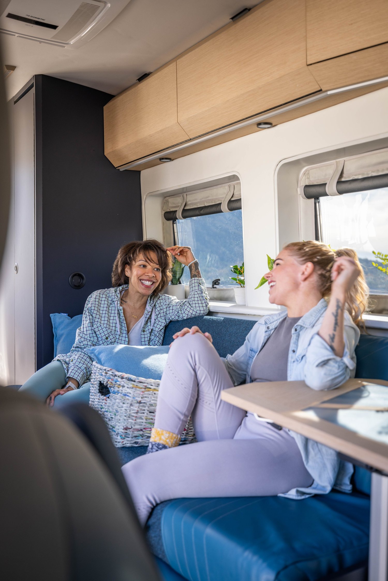 Two women laughing and talking inside a vehicle with large windows, wooden overhead storage, and blue seating.