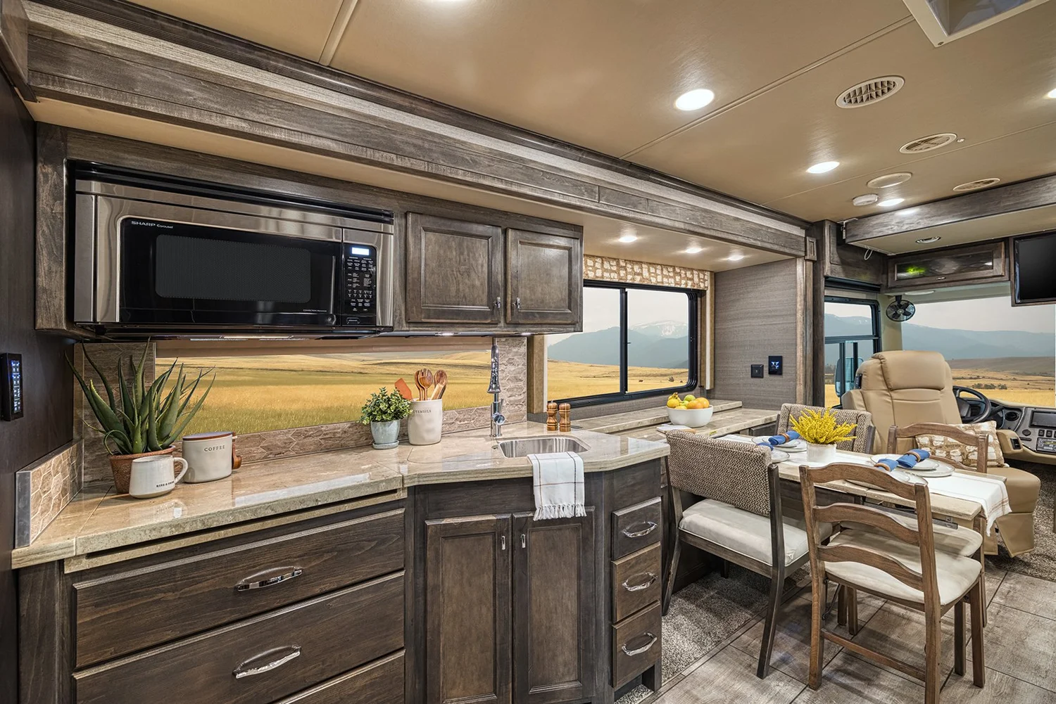 Interior of a kitchen and dining area in a recreational vehicle with a mountain and field view through the window.