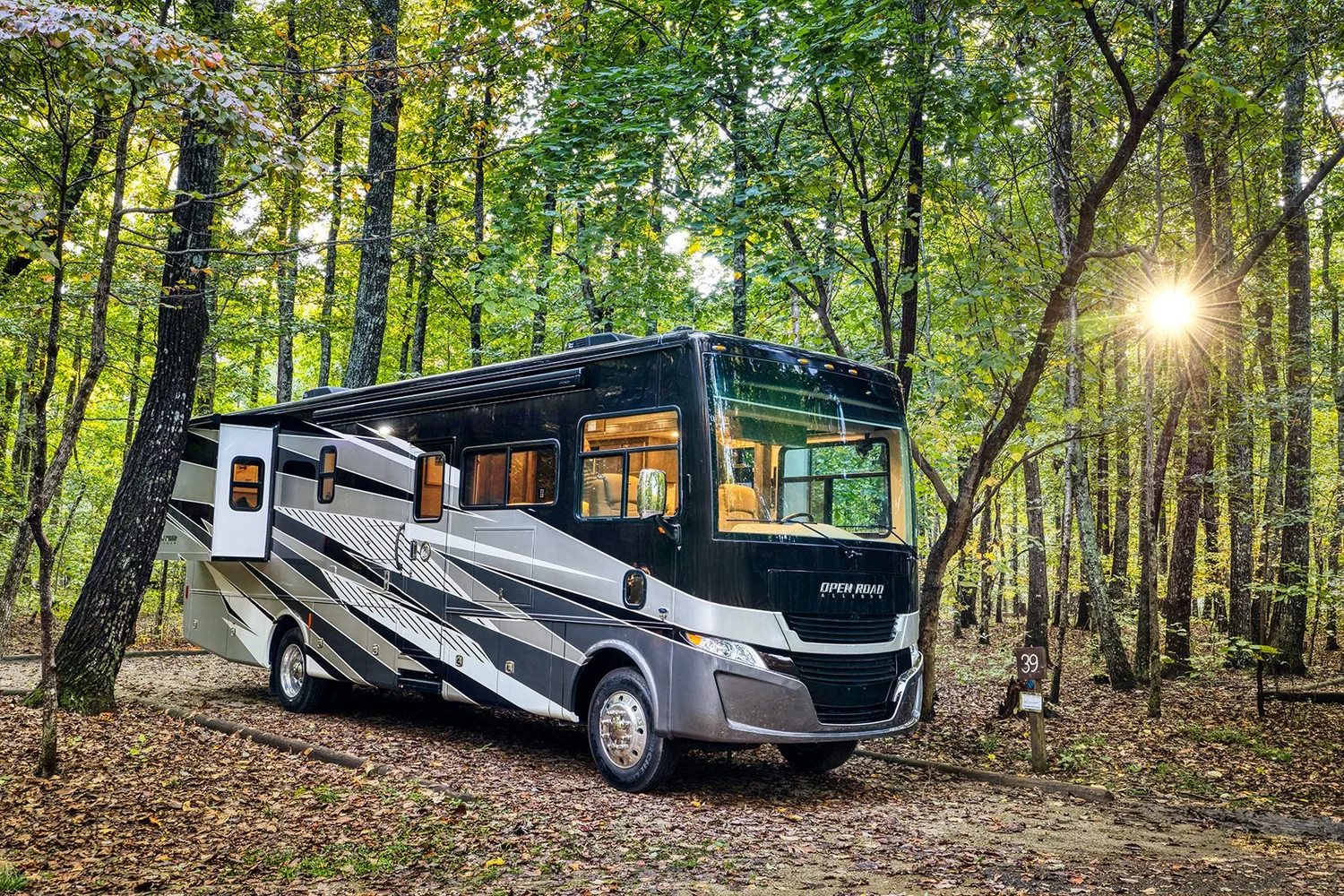 A black and white recreational vehicle (RV) parked in a forested campsite surrounded by trees with sunlight filtering through.