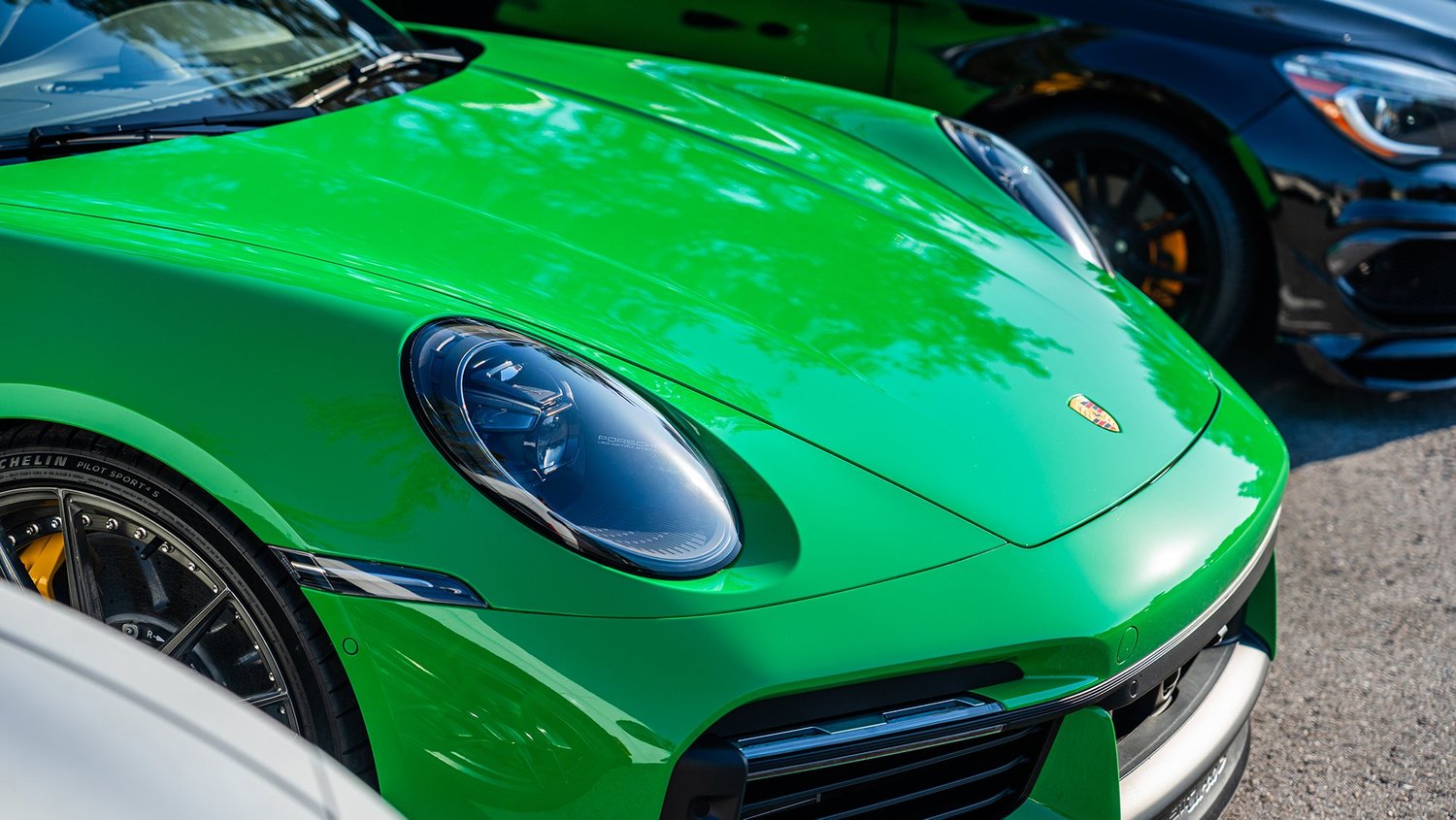 Close-up of a bright green Porsche sports car parked next to a black car, showing the front end and headlight with reflections of trees on the hood.