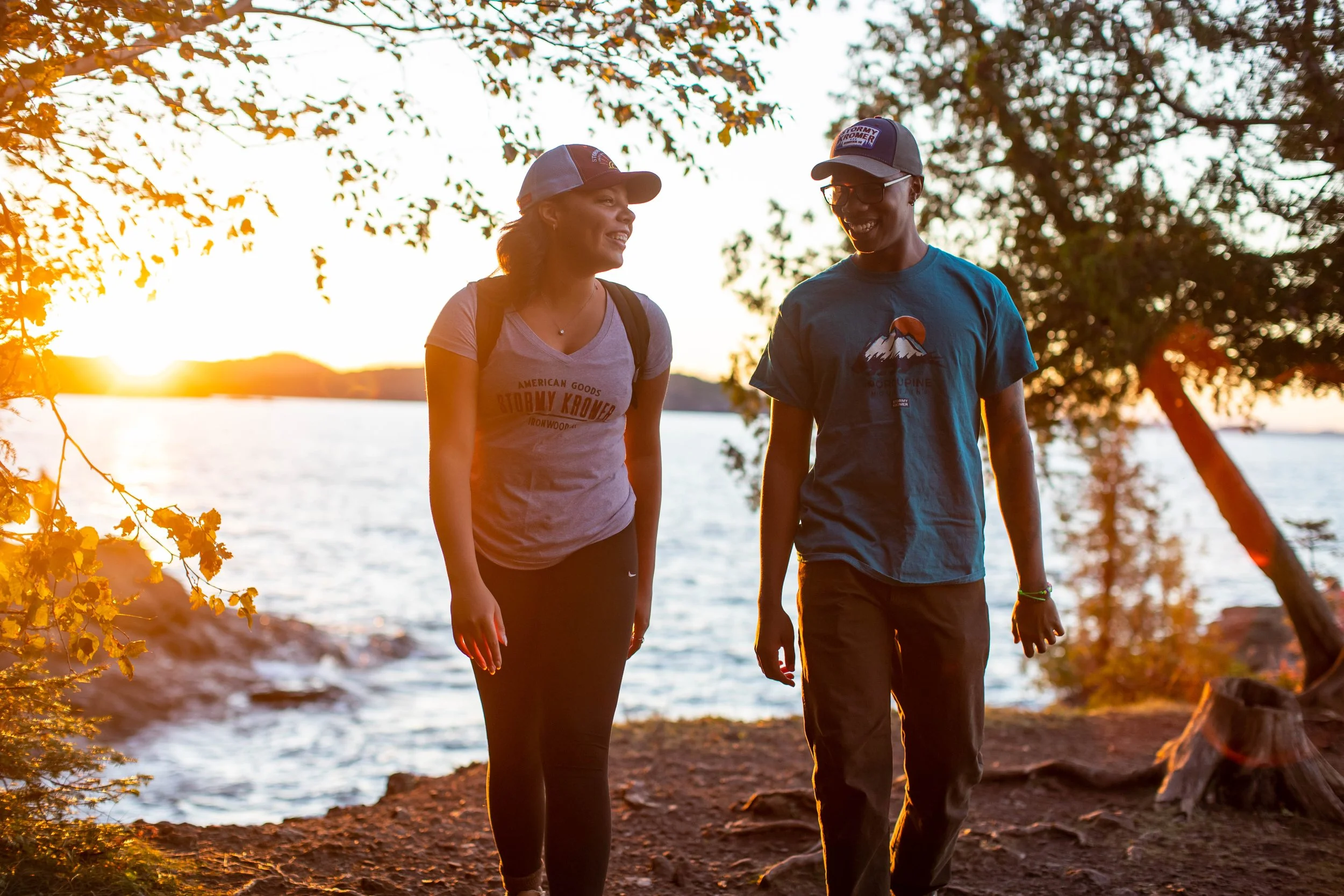 A man and woman walking and talking near a lake at sunset, surrounded by trees with orange leaves.