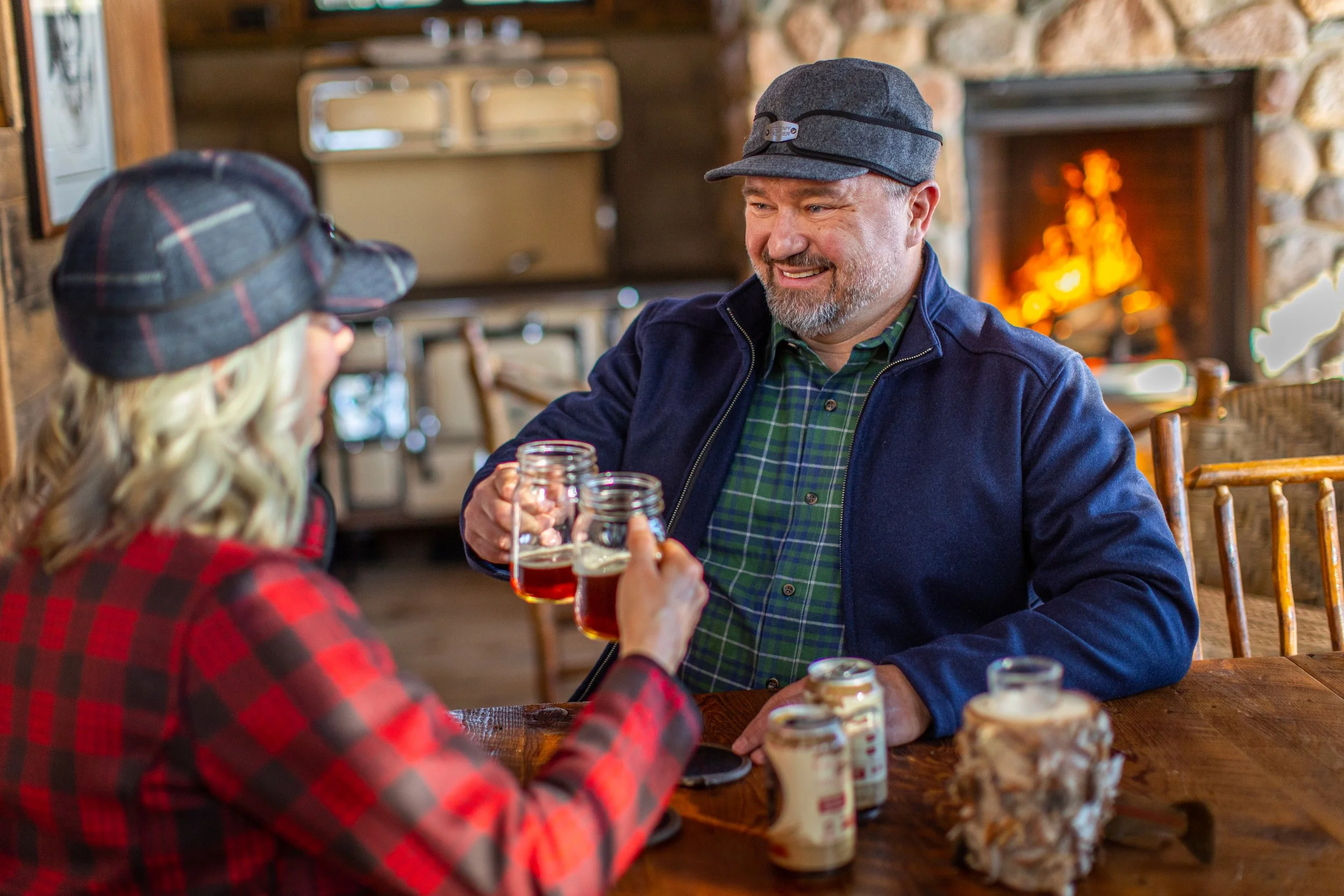 A man and a woman wearing plaid shirts and hats, sitting at a wooden table, clinking mason jars filled with a dark beverage, in a cozy cabin setting with a fireplace in the background.