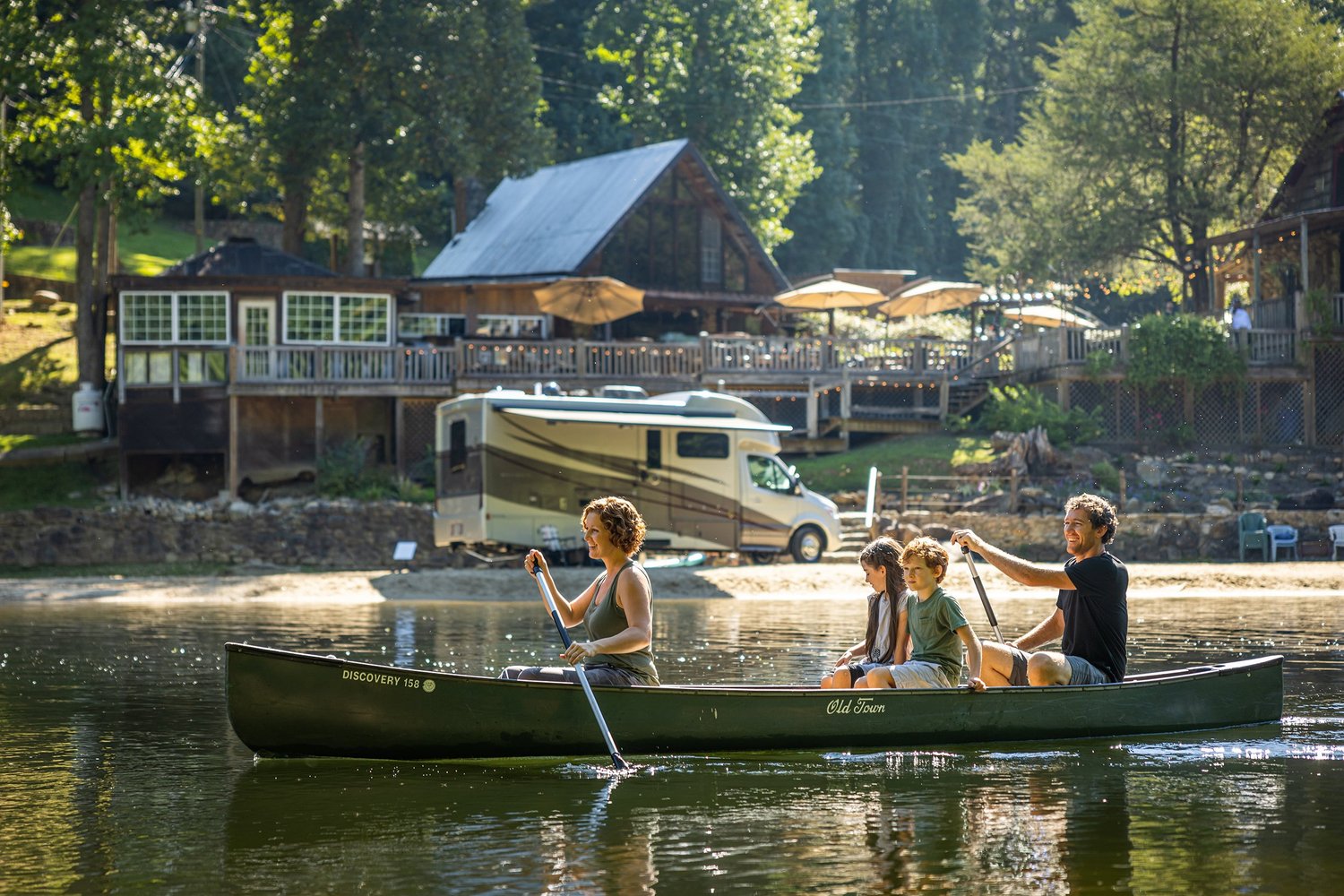 Family of four paddling a canoe on a lake with a house and trees in the background.