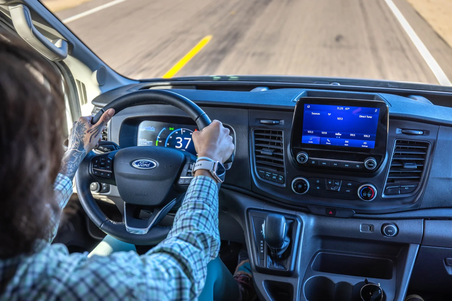 Interior of a Ford van with a person driving, hands on the steering wheel, dashboard with digital speedometer showing 37 miles per hour, and a touchscreen displaying radio options, on a highway.
