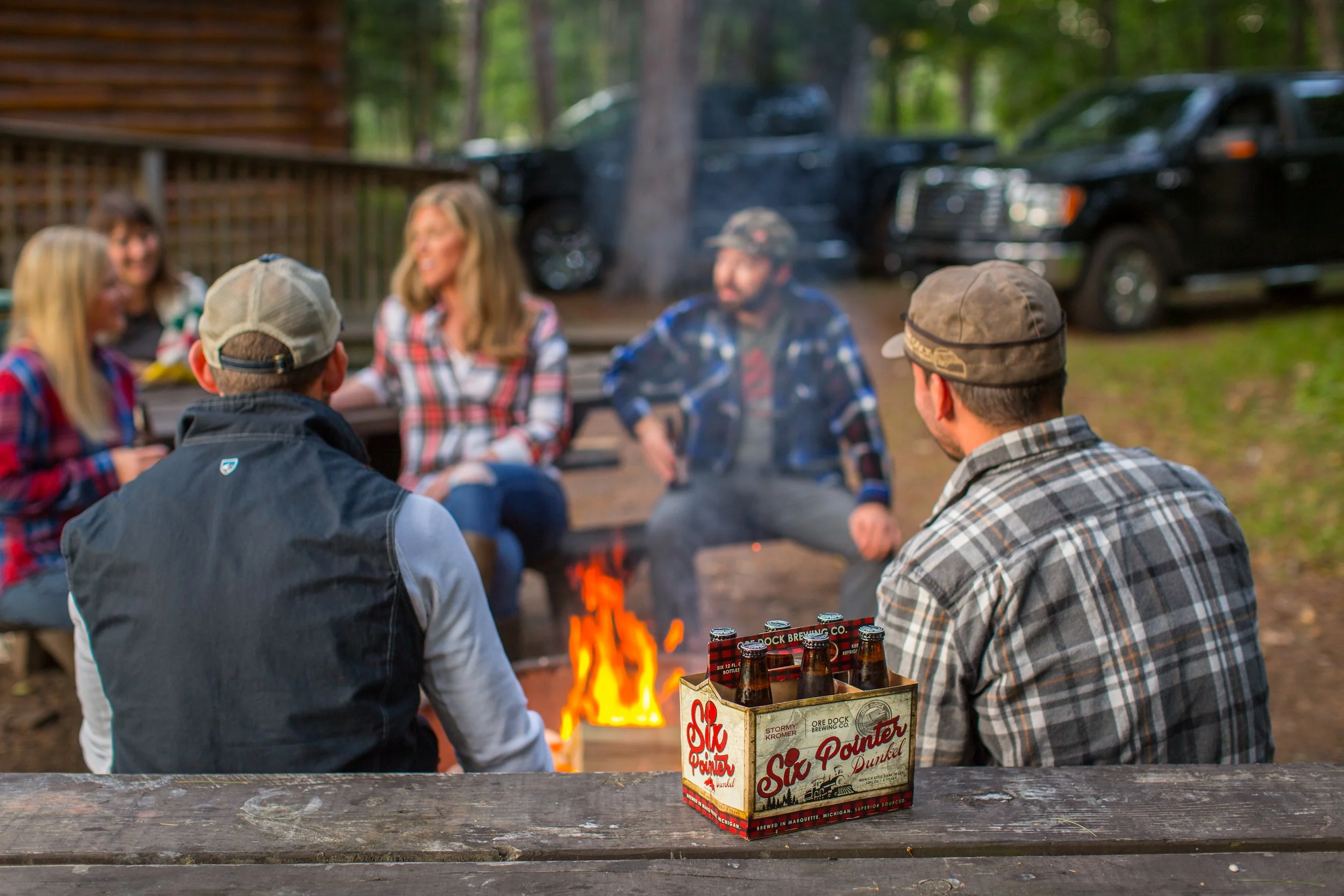 Group of friends sitting around a campfire in a wooded area with parked trucks in the background, enjoying drinks in a wooden outdoor setting.