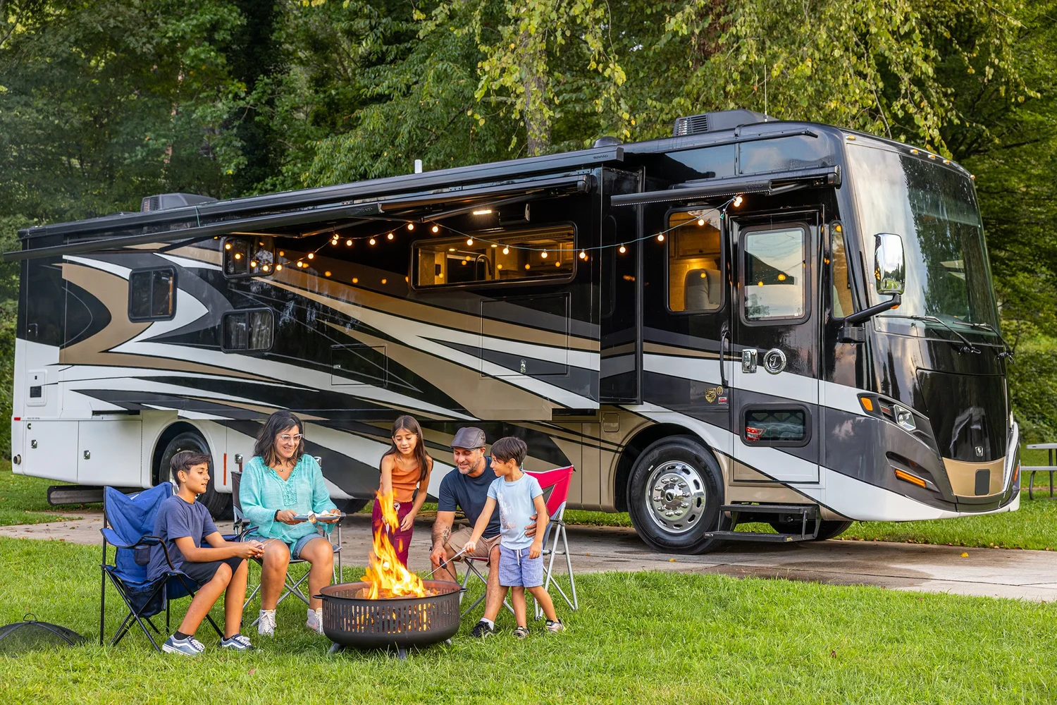 Family camping outside an RV with a fire pit, enjoying a campfire and sitting on chairs at dusk in a park with trees.