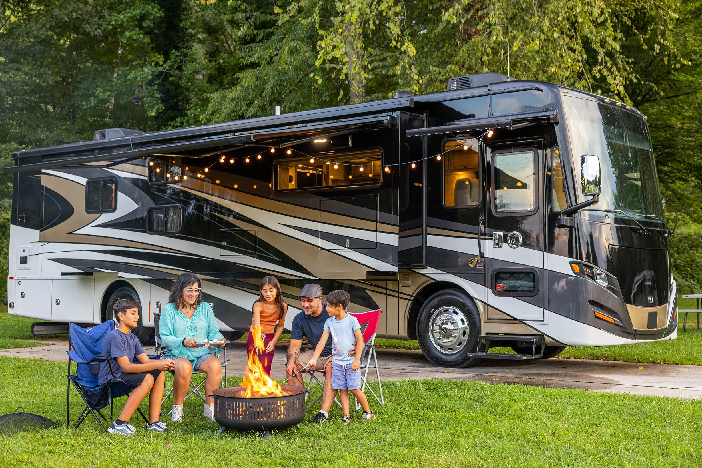 A family camping scene with five people sitting around a fire pit in front of a large black and white RV during daytime, surrounded by trees.
