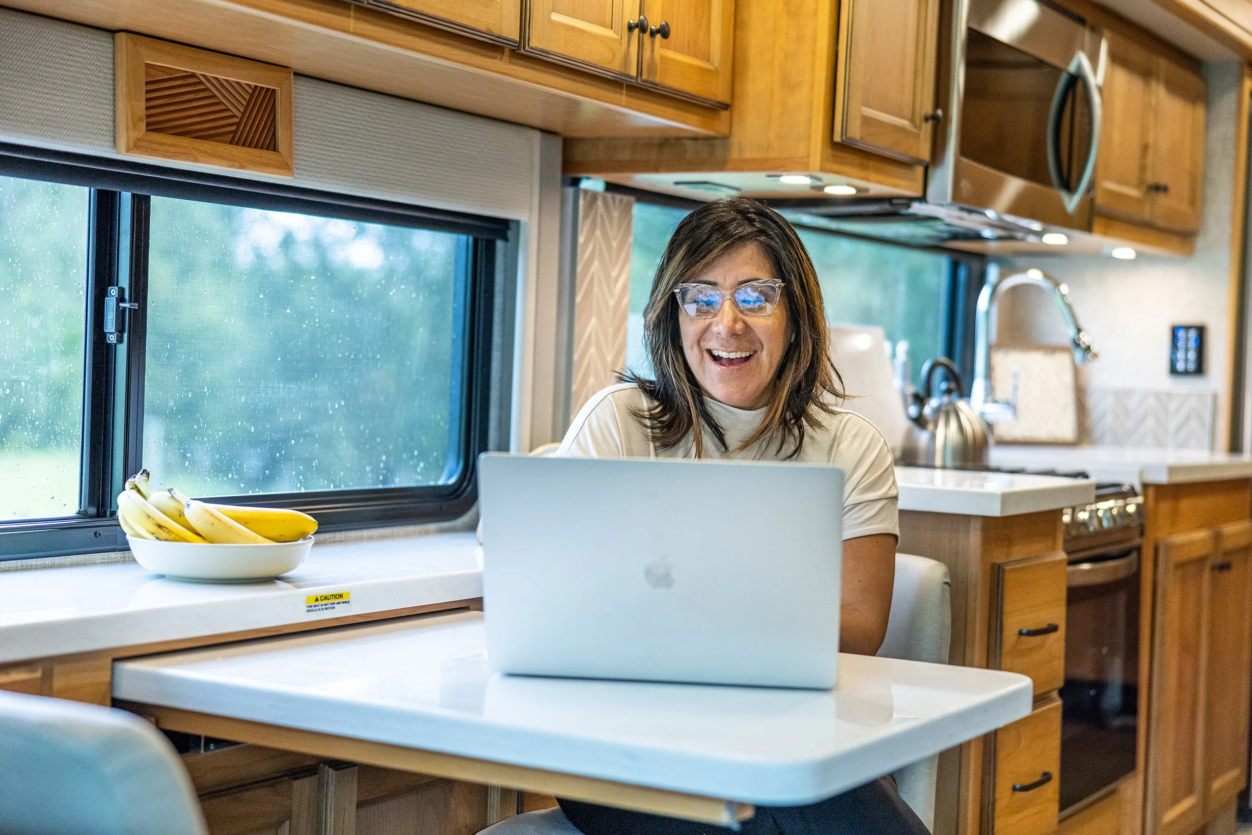 Woman with glasses smiling at laptop in kitchen with wood cabinets, bananas on table, and window showing greenery outside.