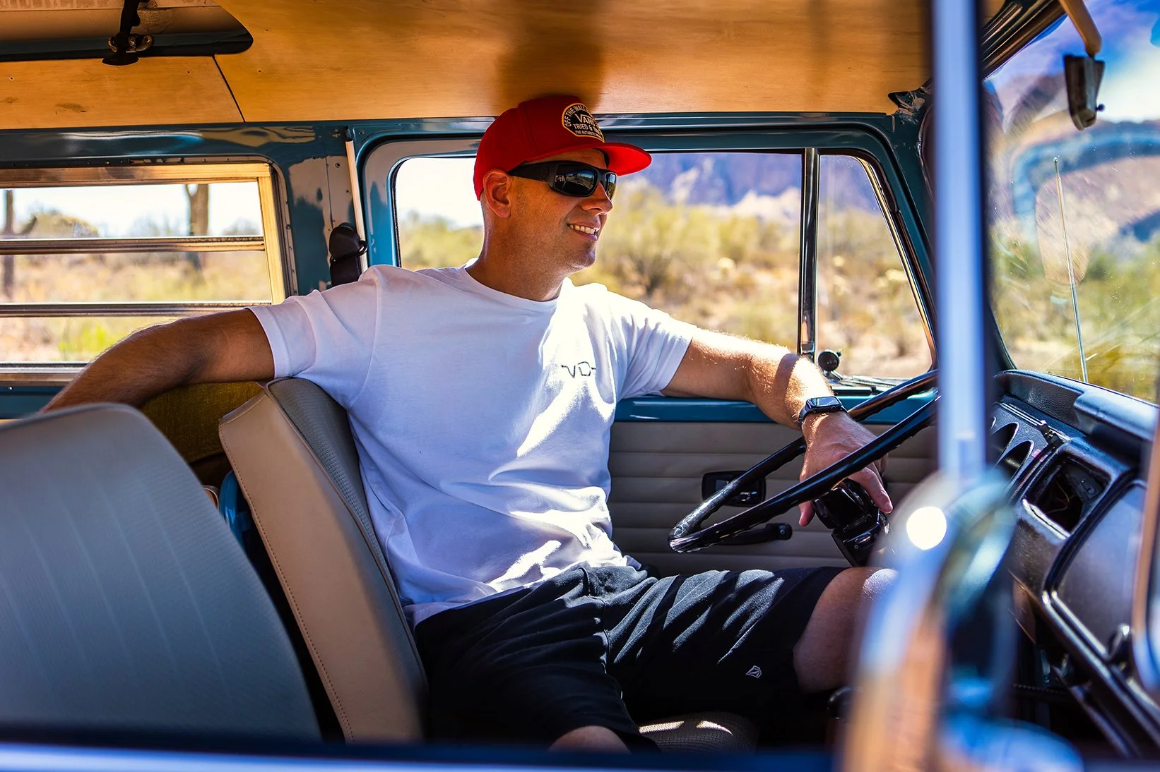 A man wearing sunglasses, a red baseball cap, a white t-shirt, and black shorts, sitting in the driver's seat of a vintage van, smiling as he looks out the window at a desert landscape with mountains in the background.
