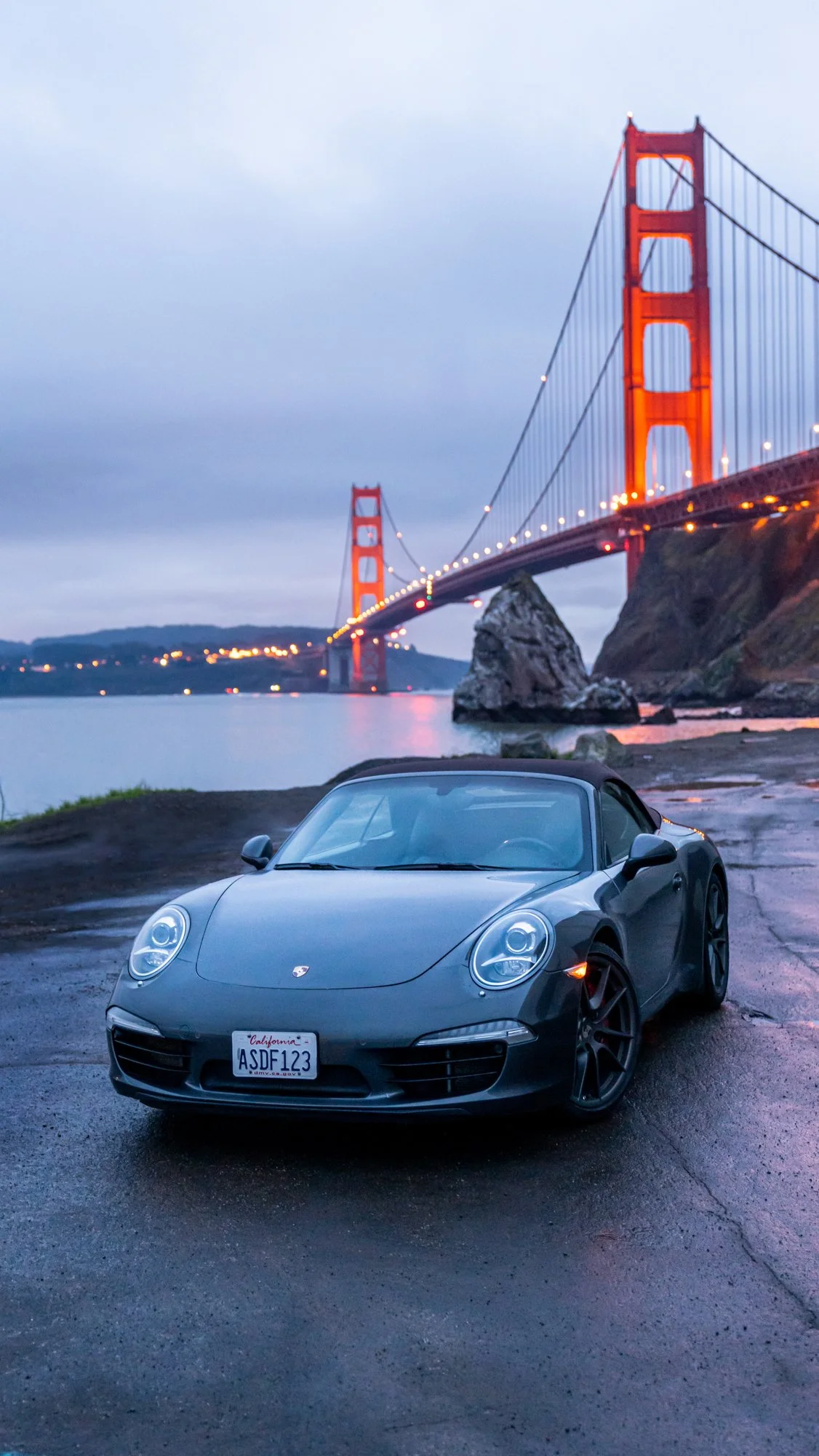 A gray Porsche convertible parked near the shoreline with the Golden Gate Bridge illuminated in the background during dusk.