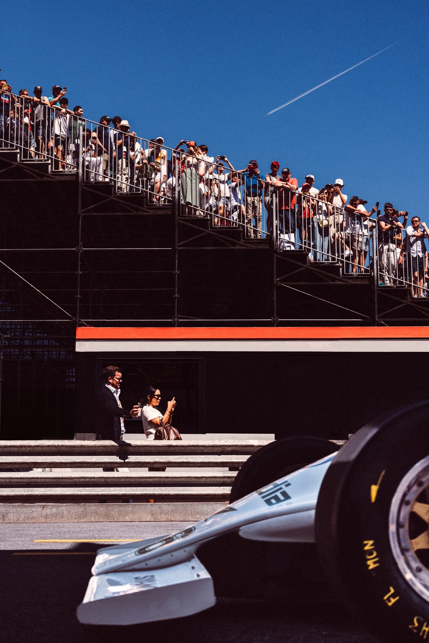 A Formula 1 race car on the track with spectators in the grandstand and people taking photos, under a clear blue sky.