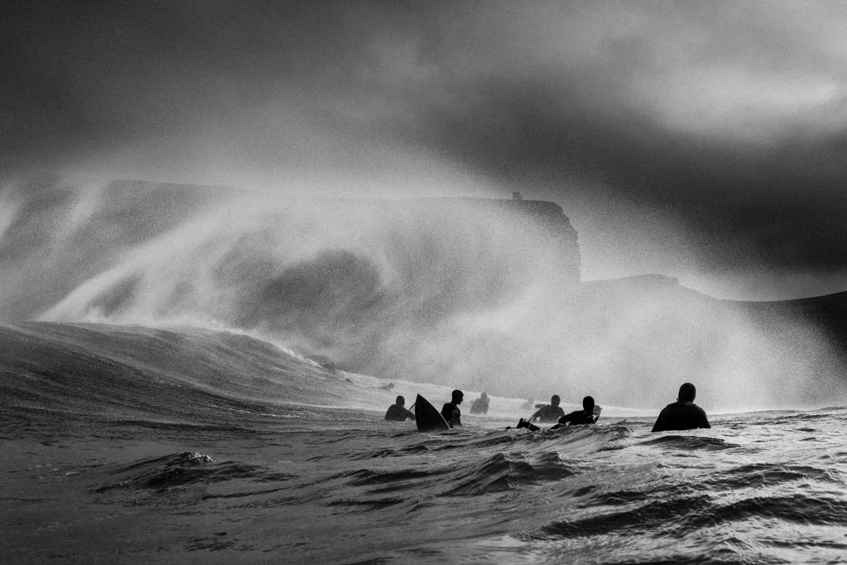 Group of surfers in the ocean with large waves and dark, cloudy sky