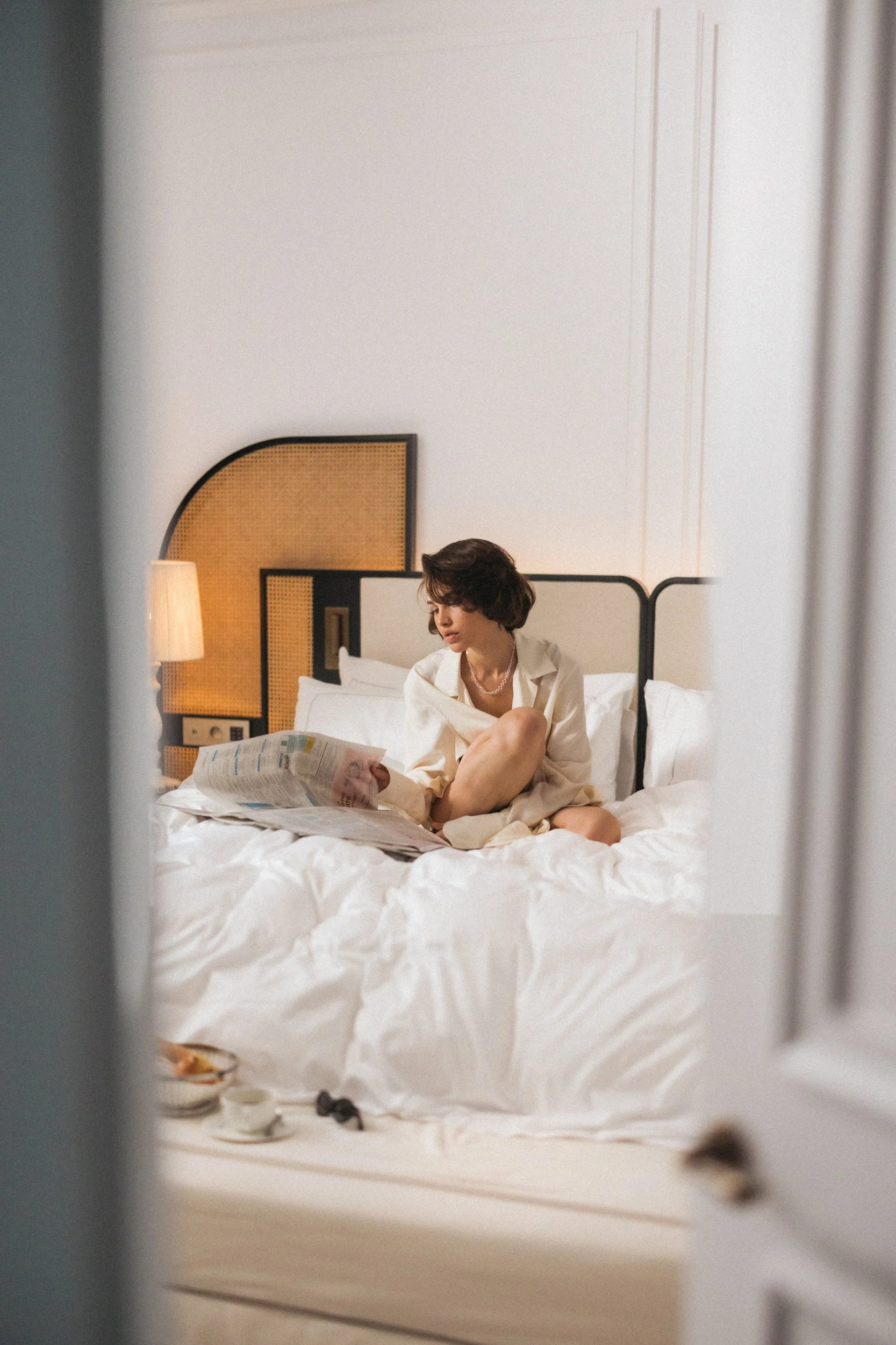 A woman sitting on a bed reading a newspaper, seen through a partially open door in a bedroom with a headboard, a lamp, and white bedding.
