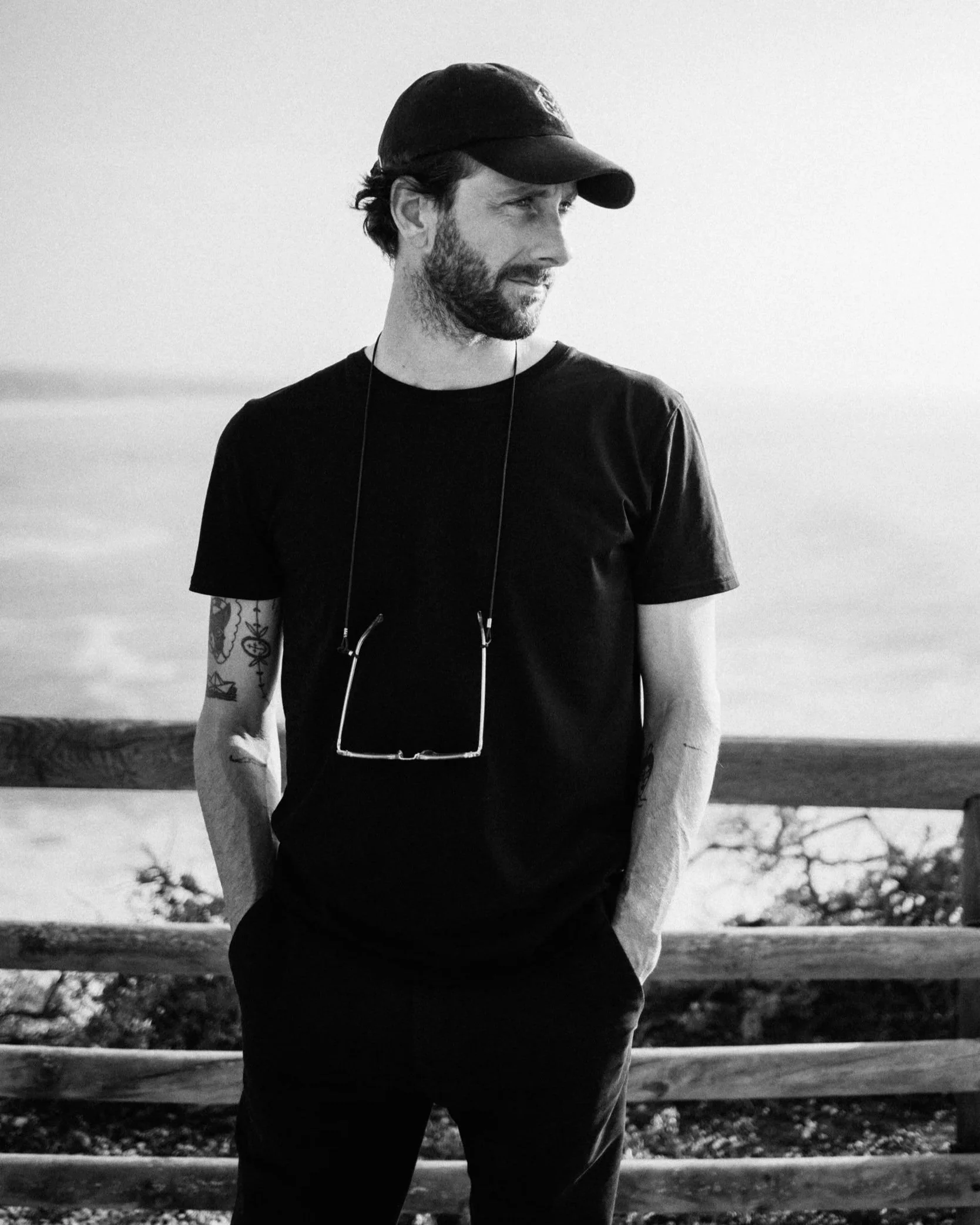 A young man with a beard and tattoos, wearing a black t-shirt, a black cap, and glasses hanging around his neck, standing outdoors by a wooden railing with water and trees in the background.