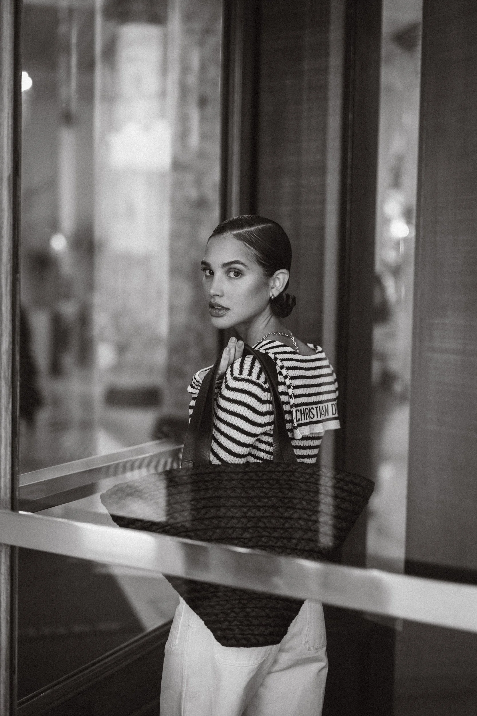 Ritz Carlton, Cannes.A woman with dark hair slicked back, wearing a striped shirt with Christian Dior, lifting her hand to her shoulder, holding a large textured bag, standing by a reflective glass door in black and white.