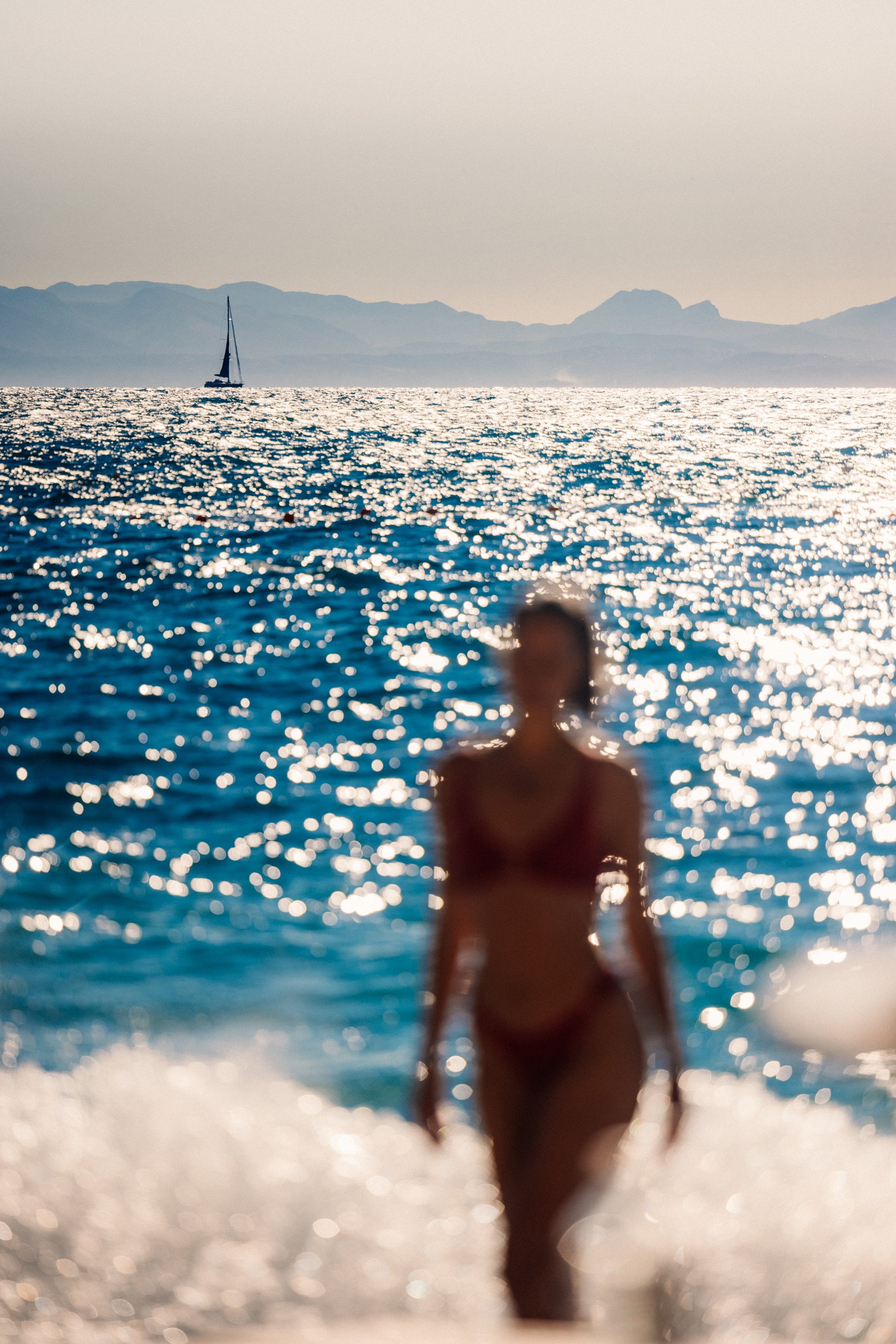A woman in a bikini walking out of the ocean with a sailboat and mountains in the background.