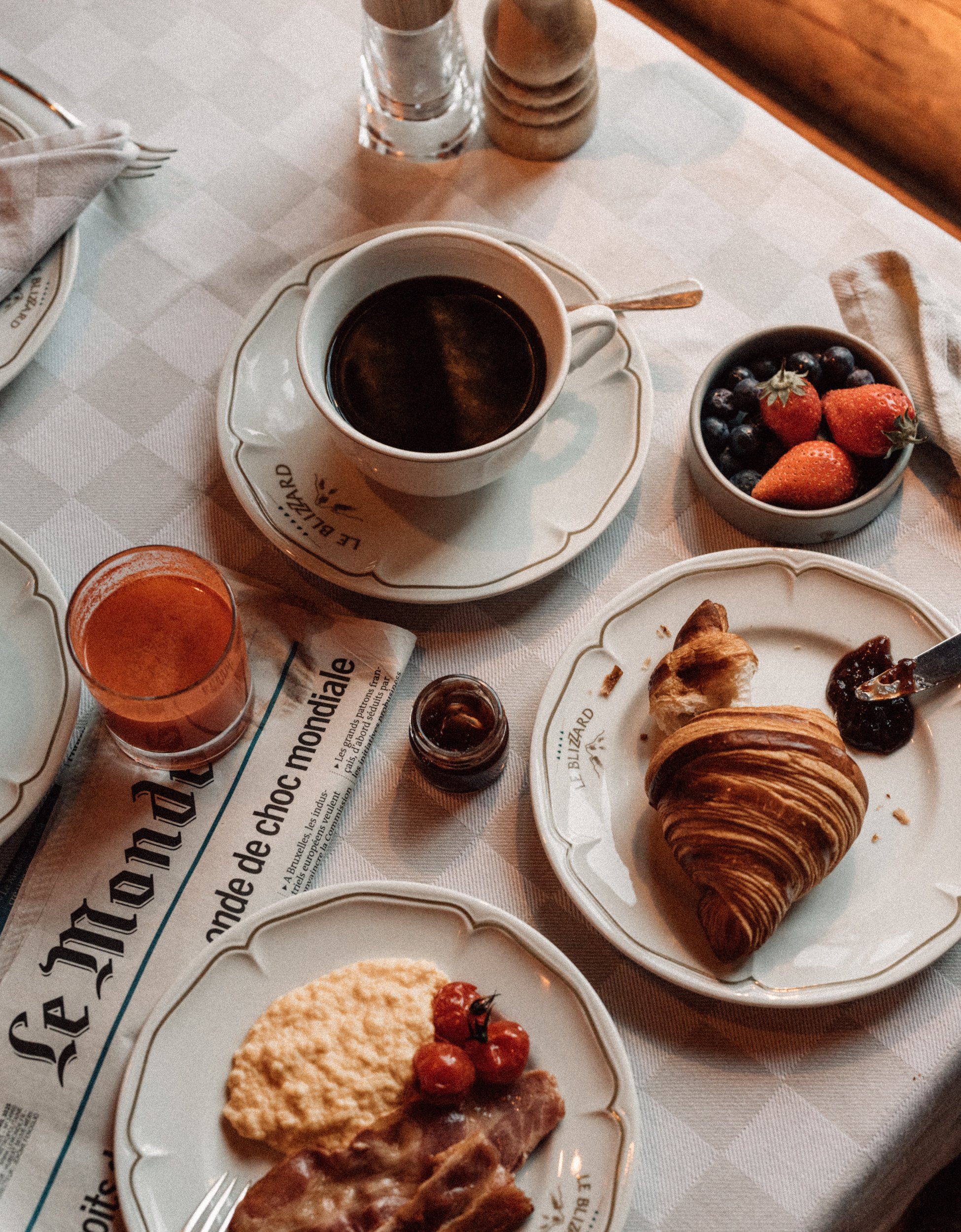 A breakfast spread on a checkered tablecloth including a cup of black coffee, a bowl of fresh strawberries and blueberries, a croissant with jam, a plate with scrambled eggs, cherry tomatoes, and bacon, a glass of orange juice, a jar of jam, and salt