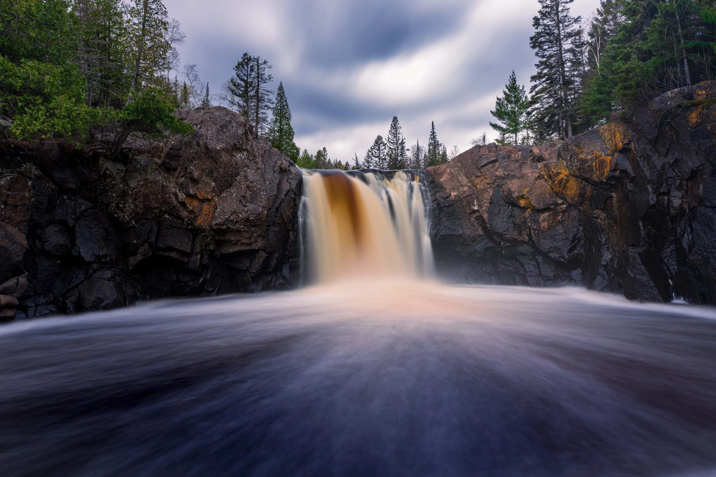 A scenic landscape of a waterfall flowing over dark rocky cliffs surrounded by evergreen trees under a cloudy sky.
