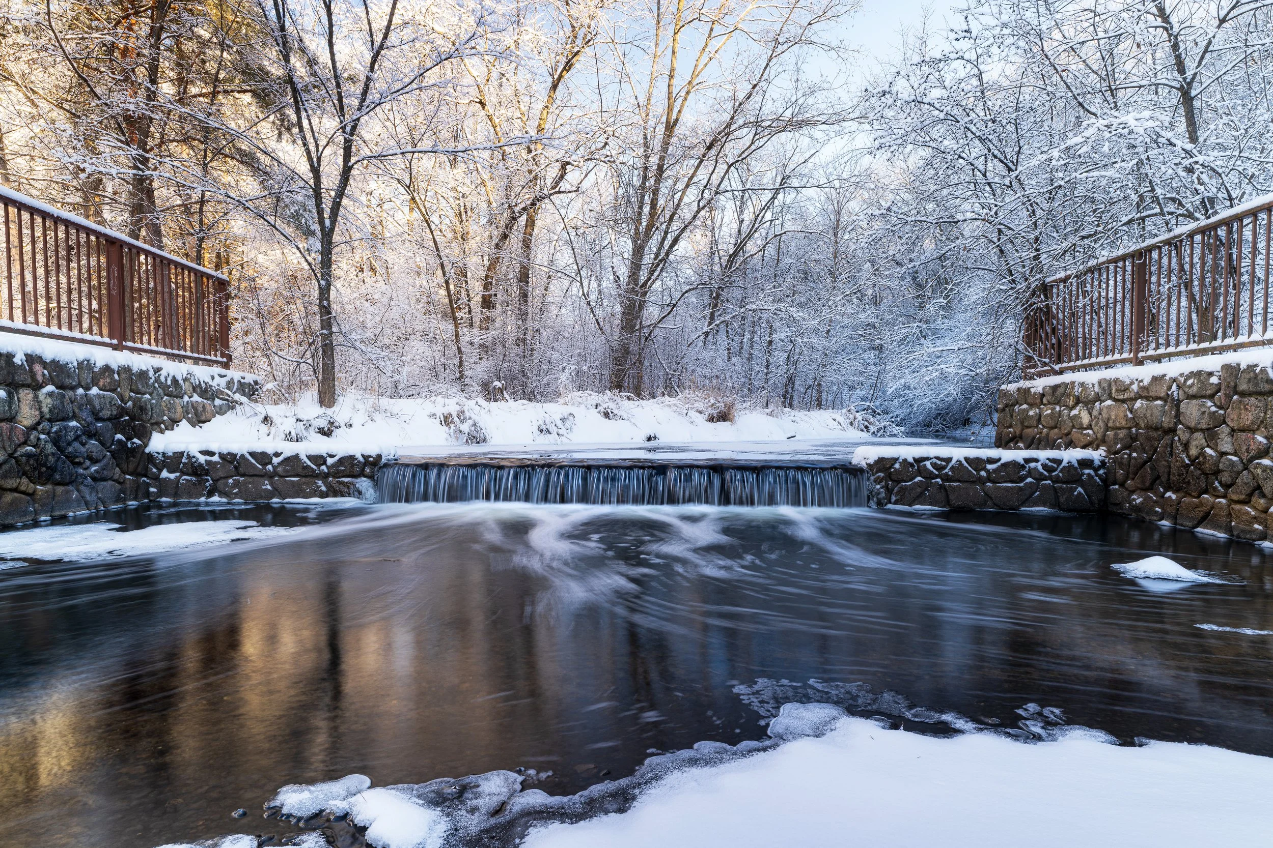 A snowy landscape with a small waterfall flowing under a bridge, surrounded by trees covered in snow.