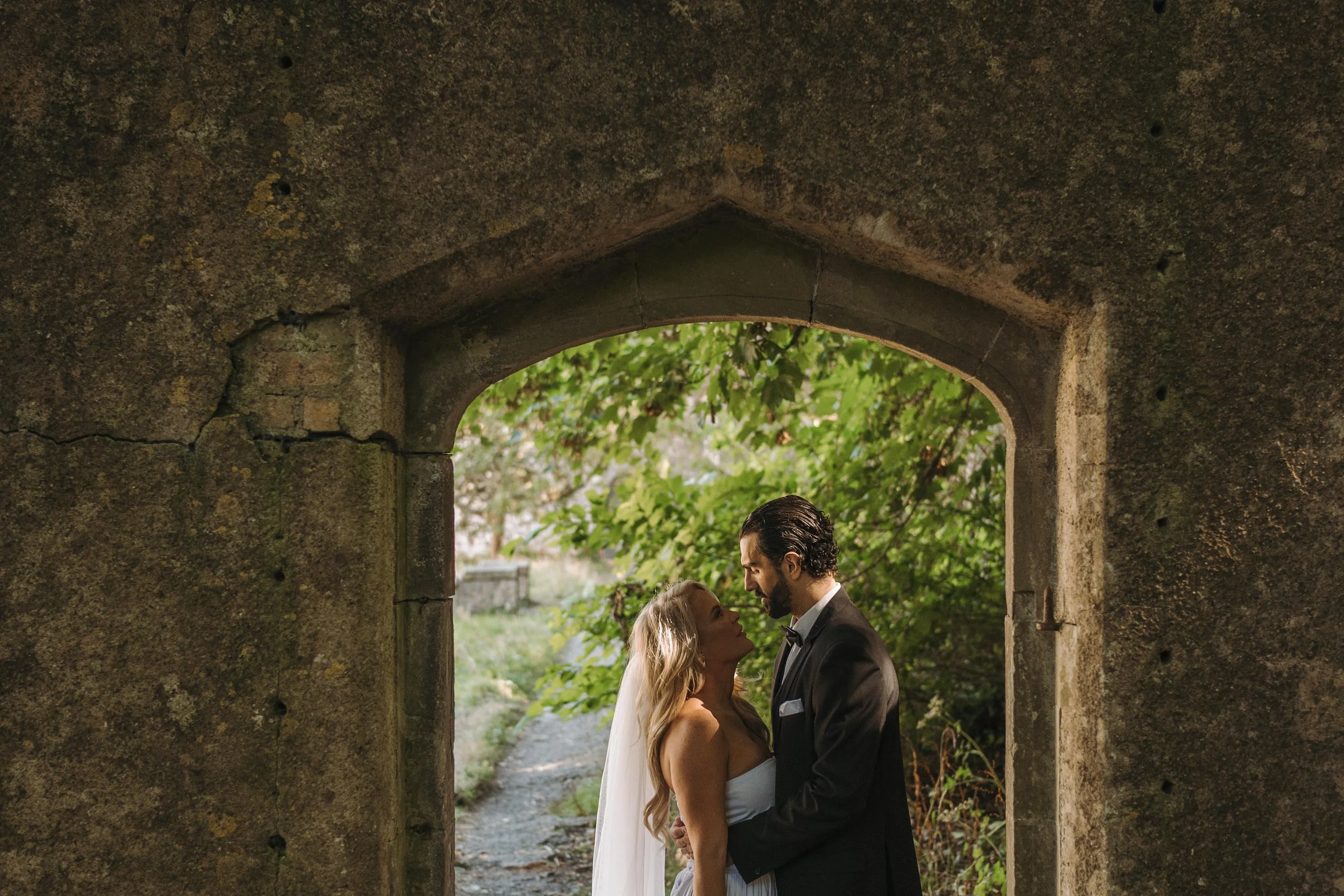 A bride and groom standing closely together under a stone archway, looking into each other's eyes surrounded by greenery.