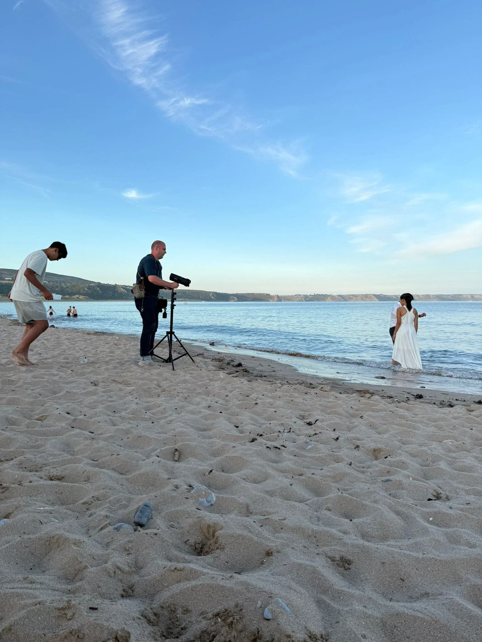 A woman in a white dress standing in the water at the beach with a man behind her, while a crew member films with a camera on a tripod and another person walks on the sand.