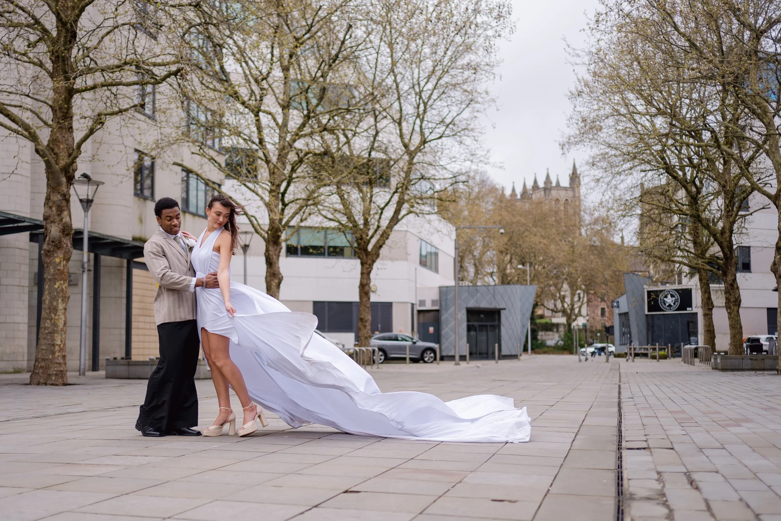 A couple dancing outdoors in formal attire, with the woman in a white gown and the man in a tan suit, on a paved city square surrounded by leafless trees and modern buildings.
