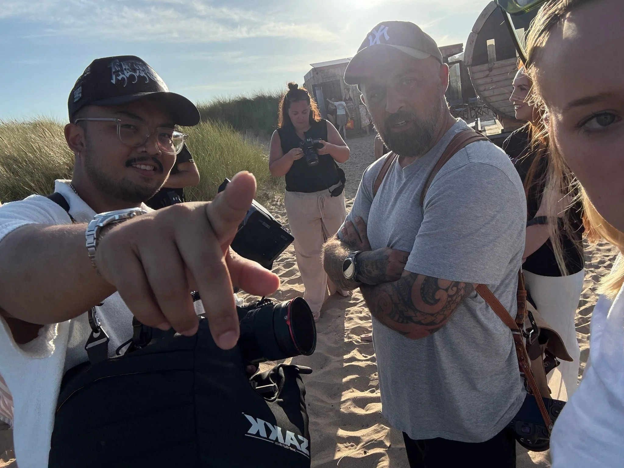 Group of people on a beach in the late afternoon, taking a selfie and looking at the camera, with grassy dunes and small beach huts in the background.