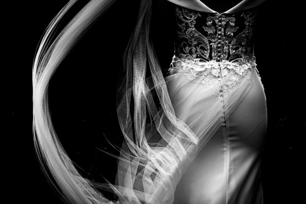 Close-up of a bride's wedding gown with lace and embroidery, with flowing fabric and a long veil against a black background.